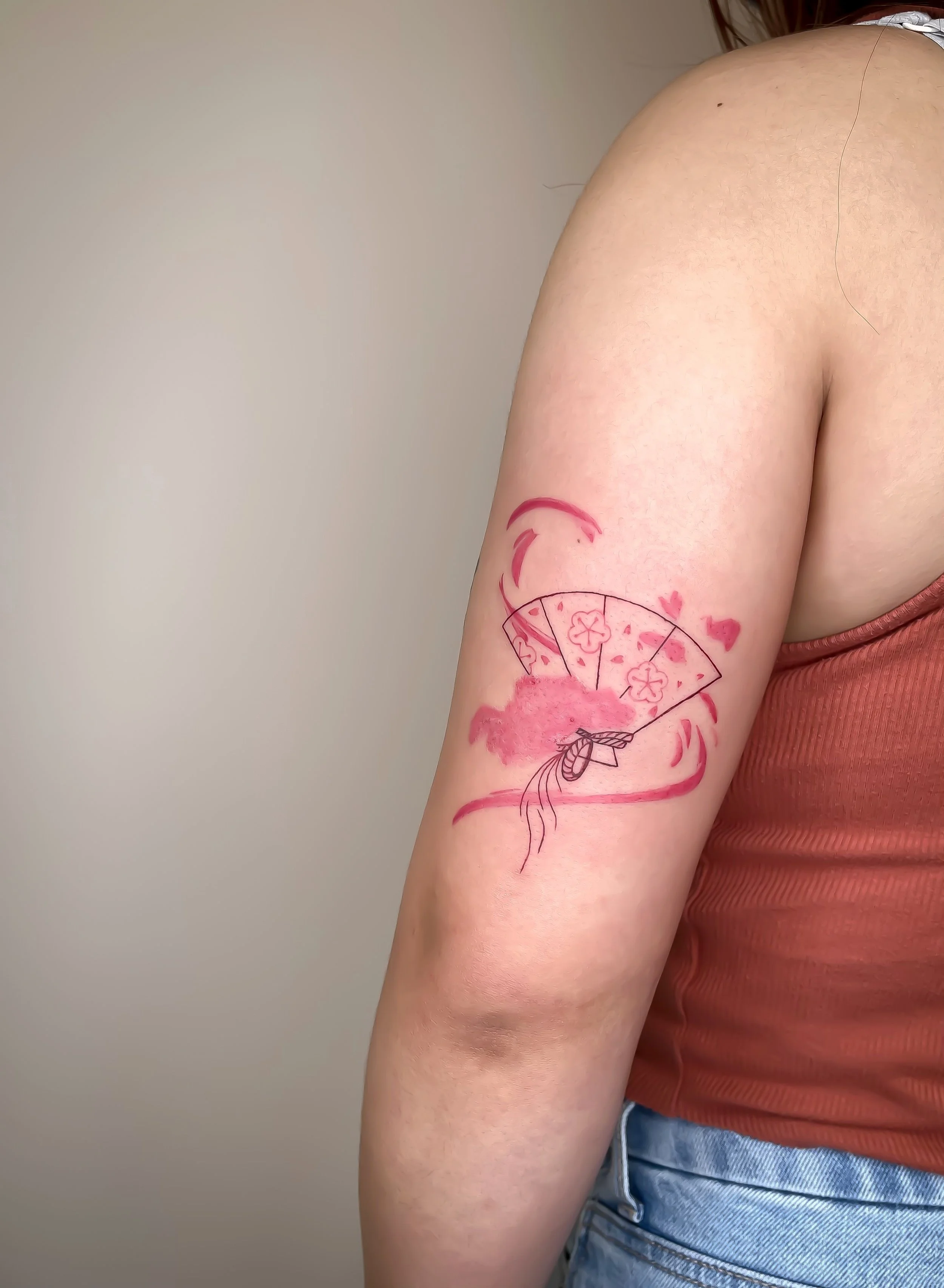 Tattoo of a pink hand fan with cherry blossoms, clouds, and hearts on a person's upper arm.