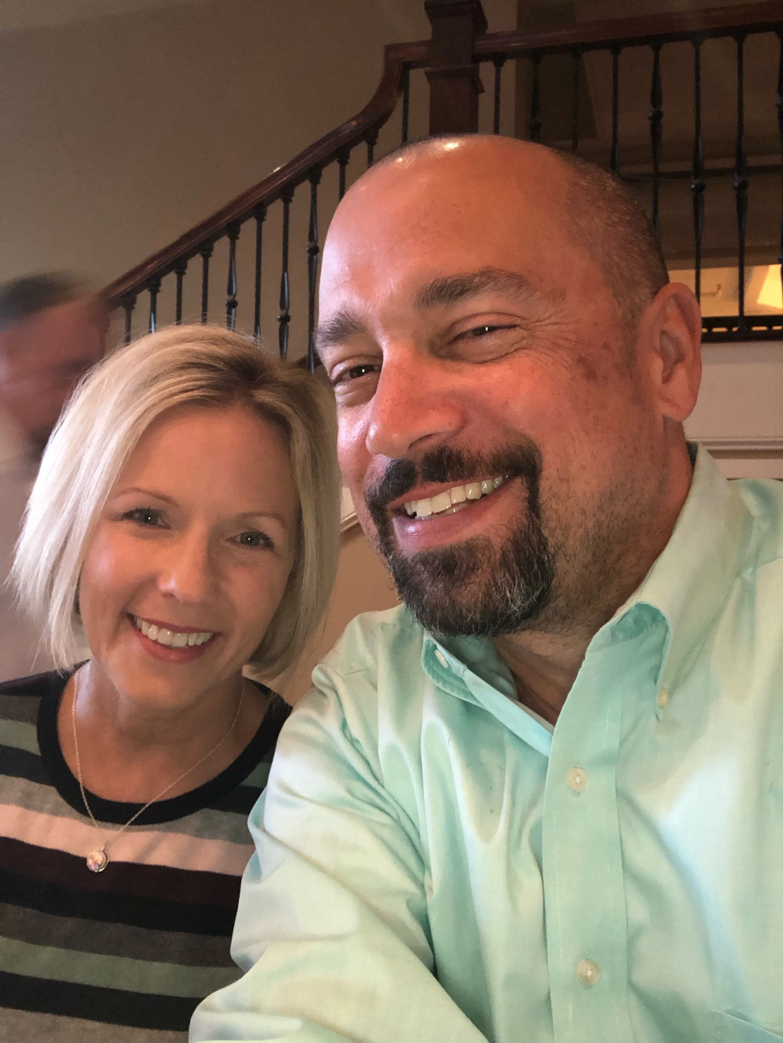 A smiling woman with blonde hair and a man with a beard taking a selfie together indoors, with a staircase in the background.