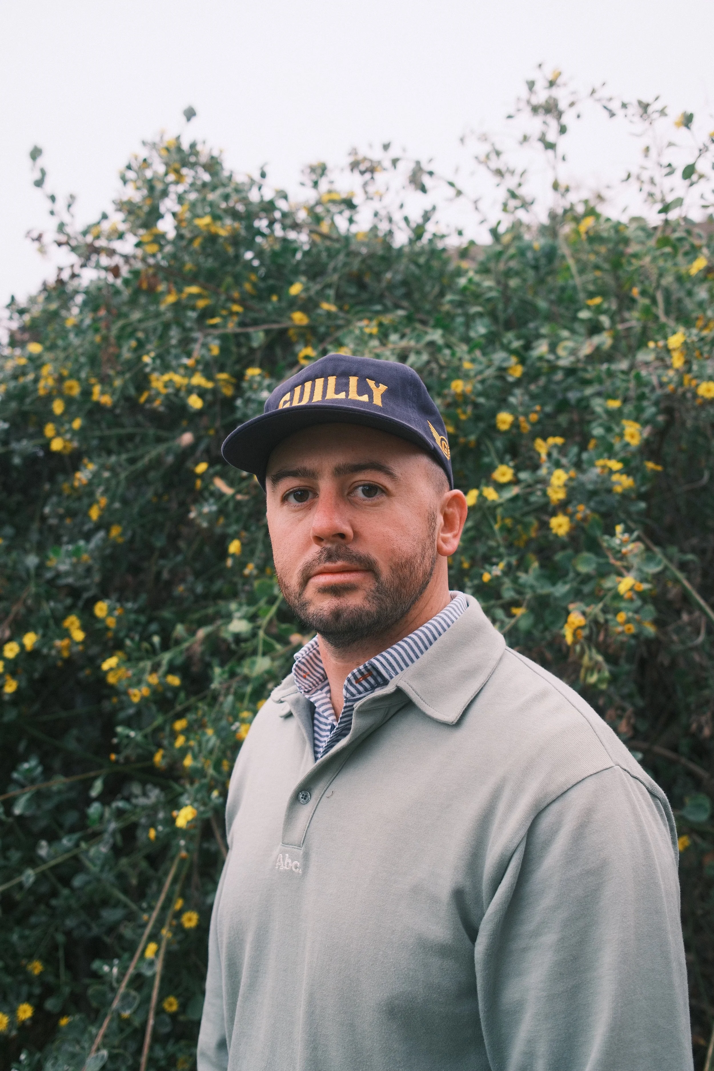 Man with beard wearing a navy blue baseball cap and a teal polo standing in front of yellow and green flowers
