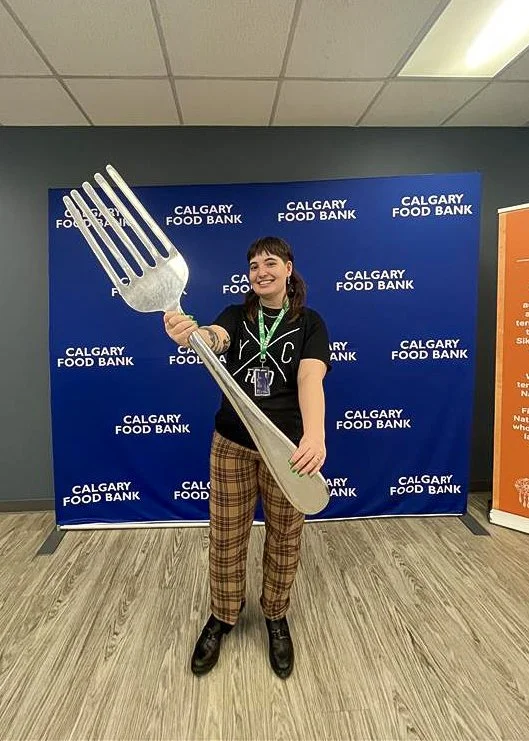 Joelle standing in front of a Calgary Food Bank backdrop holding an oversized fork.