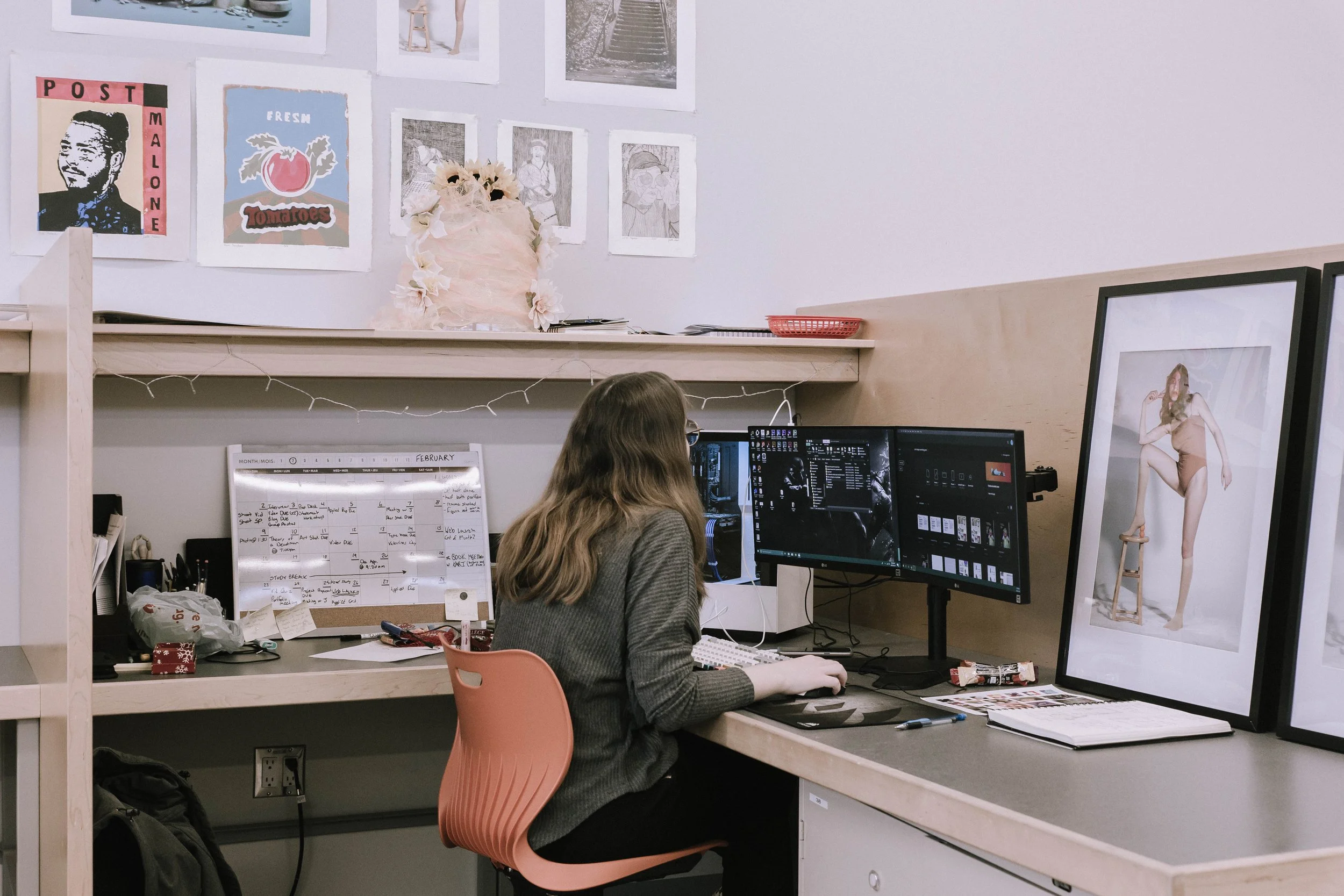 Joelle sitting at a desk working on multiple monitors in an office, with framed photographs and artwork on the wall and a whiteboard with a schedule.