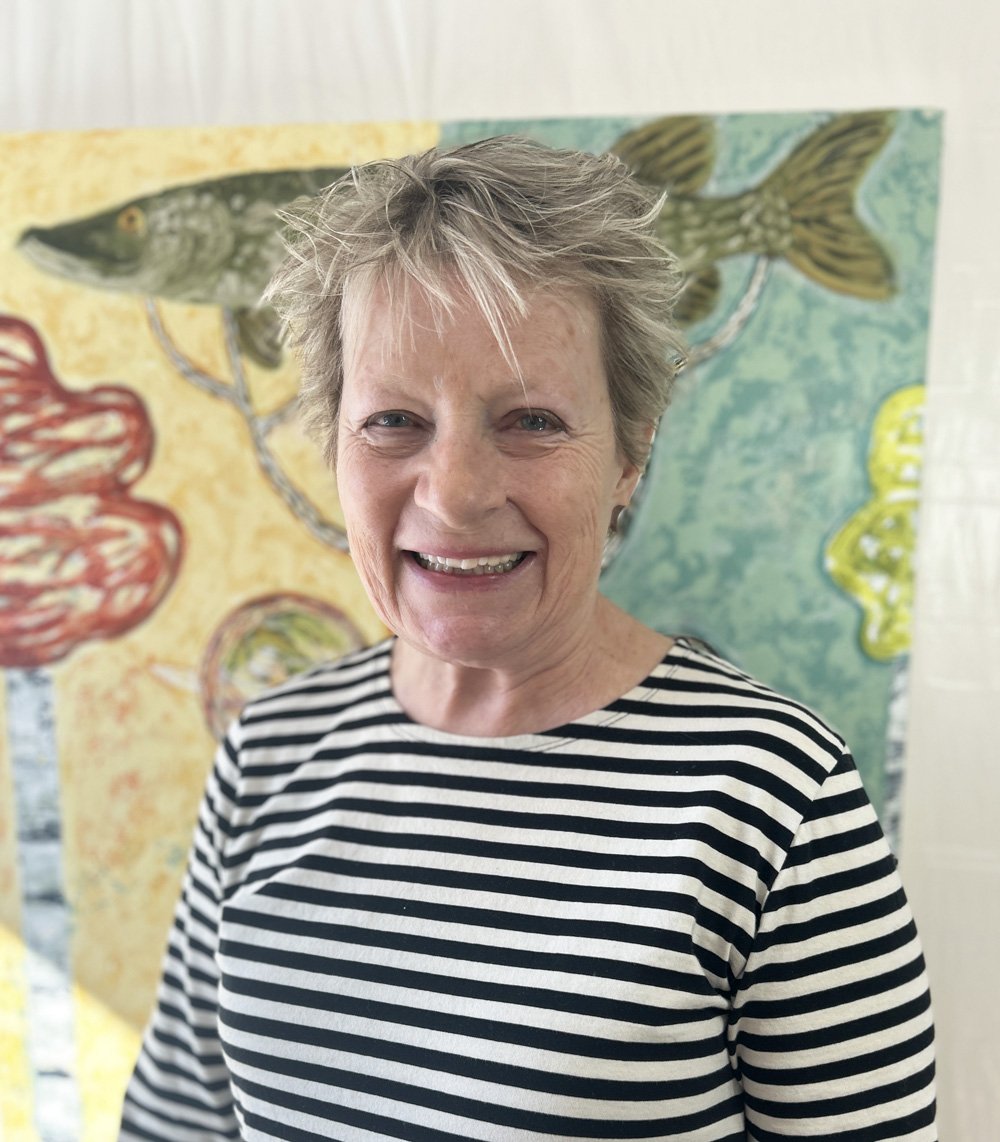 Caroline Anderson stands, smiling, in front of the painting "Northern Pike"