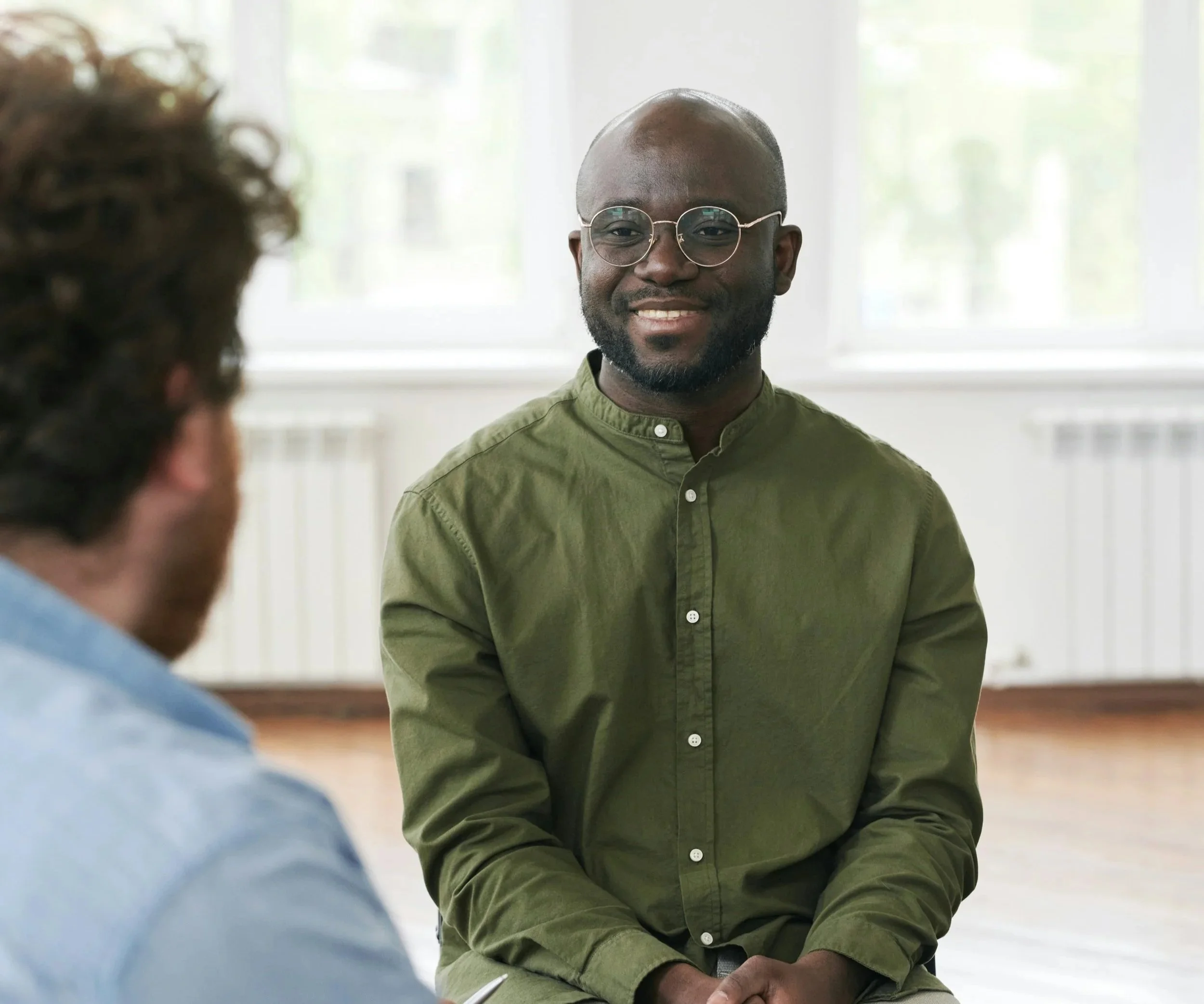 A man with glasses and a beard smiling during a conversation in a bright room with large windows.