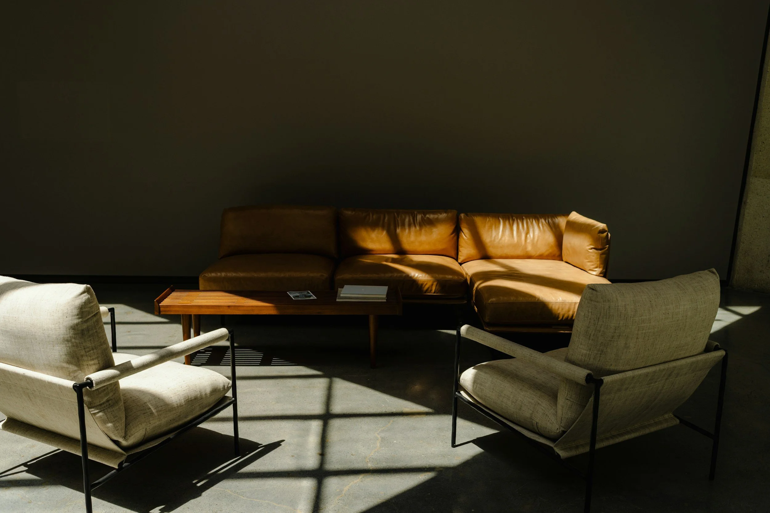 A brown leather sofa with three cushions is positioned against a plain dark wall. Two beige armchairs with black metallic frames are in the foreground facing the sofa. A wooden coffee table with some items on it is in front of the sofa, and sunlight creates shadows on the floor.
