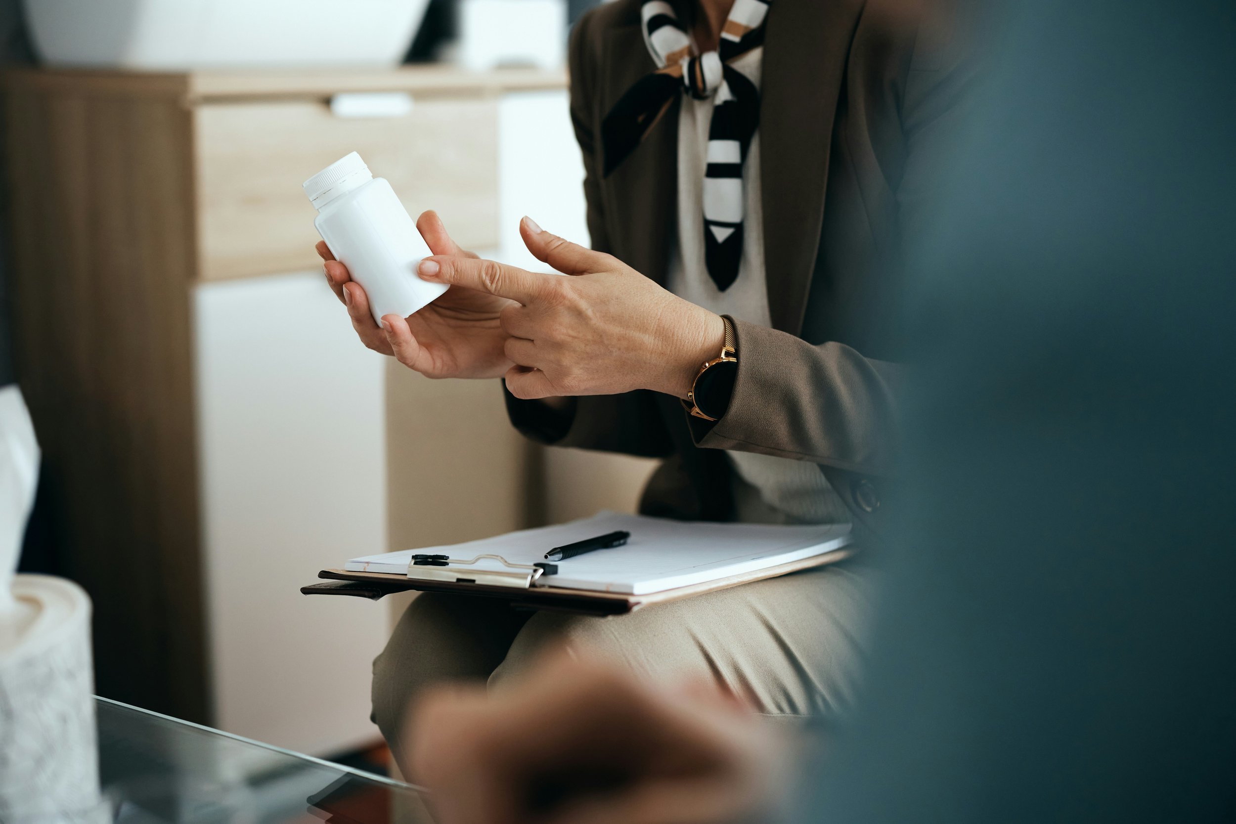 A person holding a white medication bottle in their hand, seated at a desk with a clipboard, pen, and some papers, in an indoor setting.