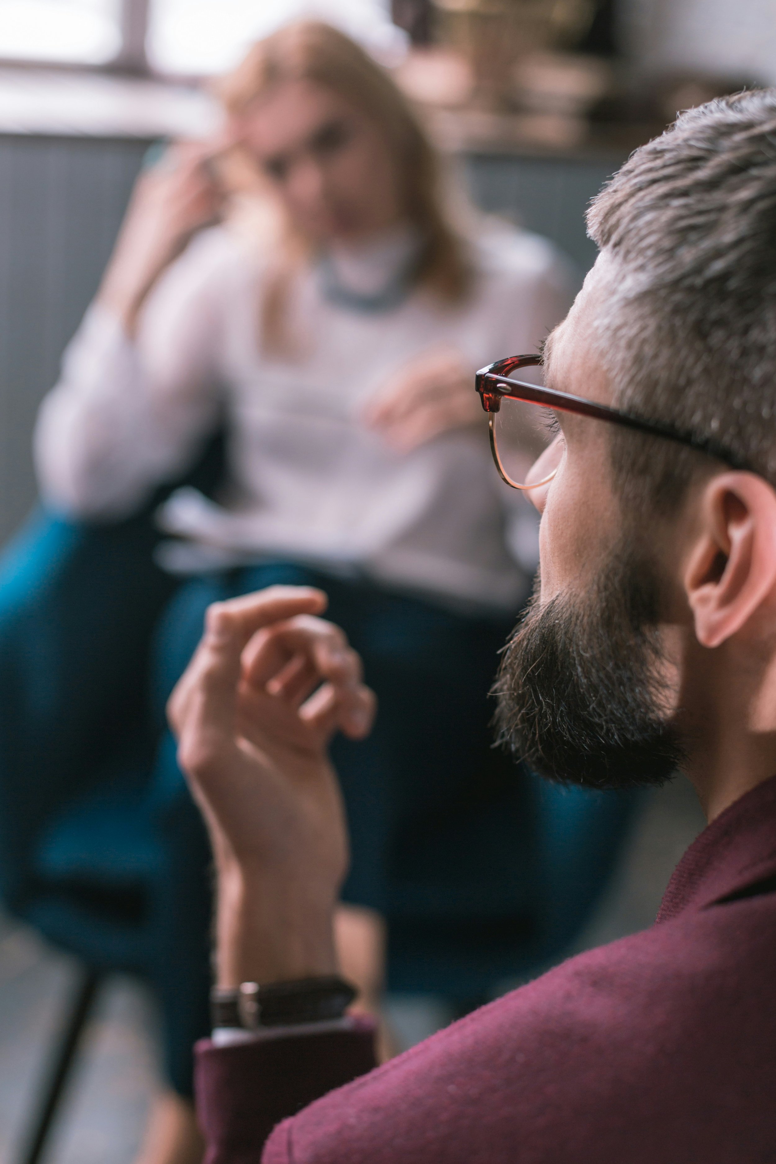 A man in glasses has his eyes closed, sitting with his head tilted back, while a woman in the background, slightly blurred, is blurred and appears to be talking or listening.