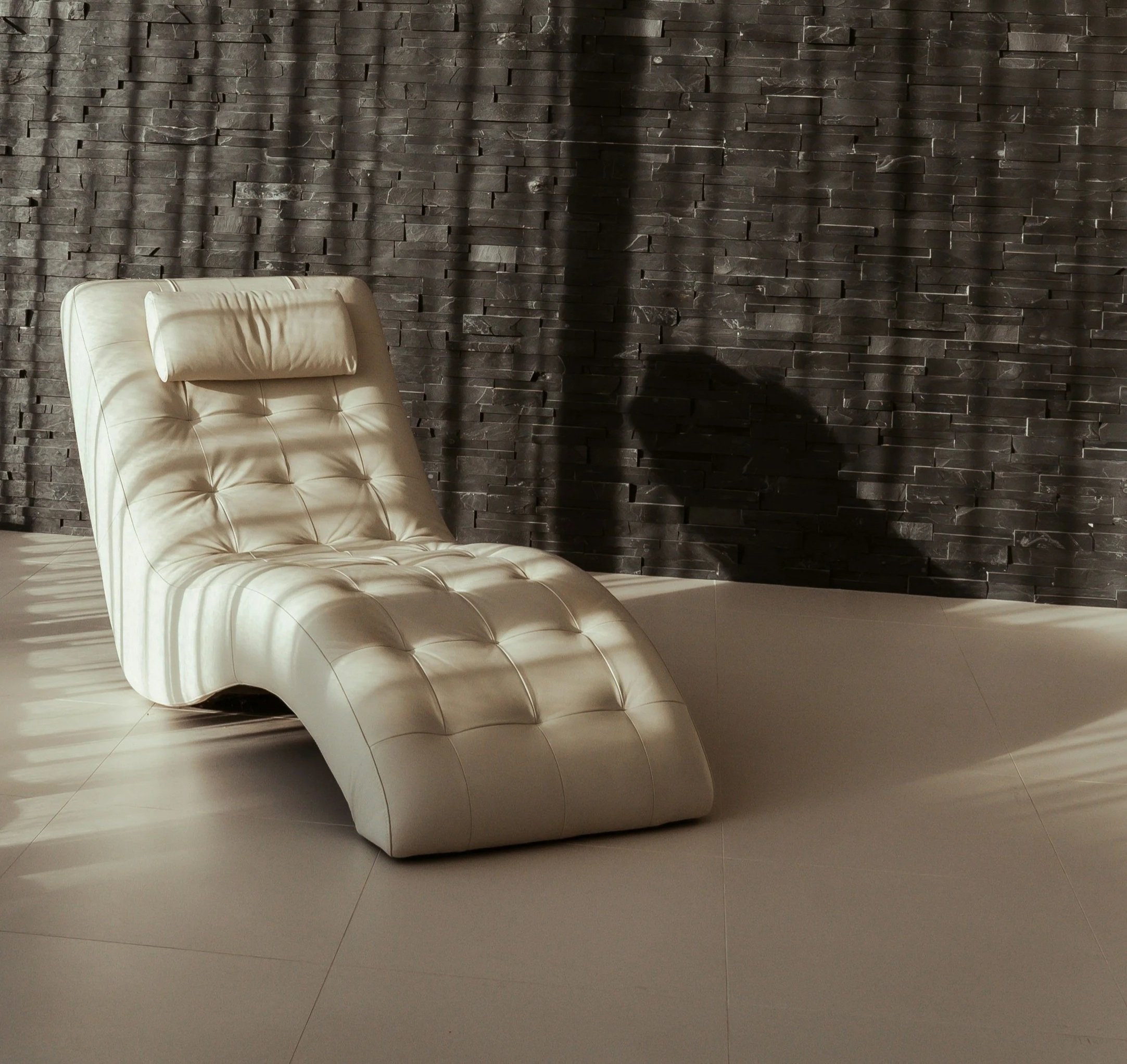 A modern lounge chair with a tufted design, made of light-colored leather, positioned on a tiled floor in front of a textured dark stone wall. Shadows cast by window blinds create striped patterns on the chair and the floor.