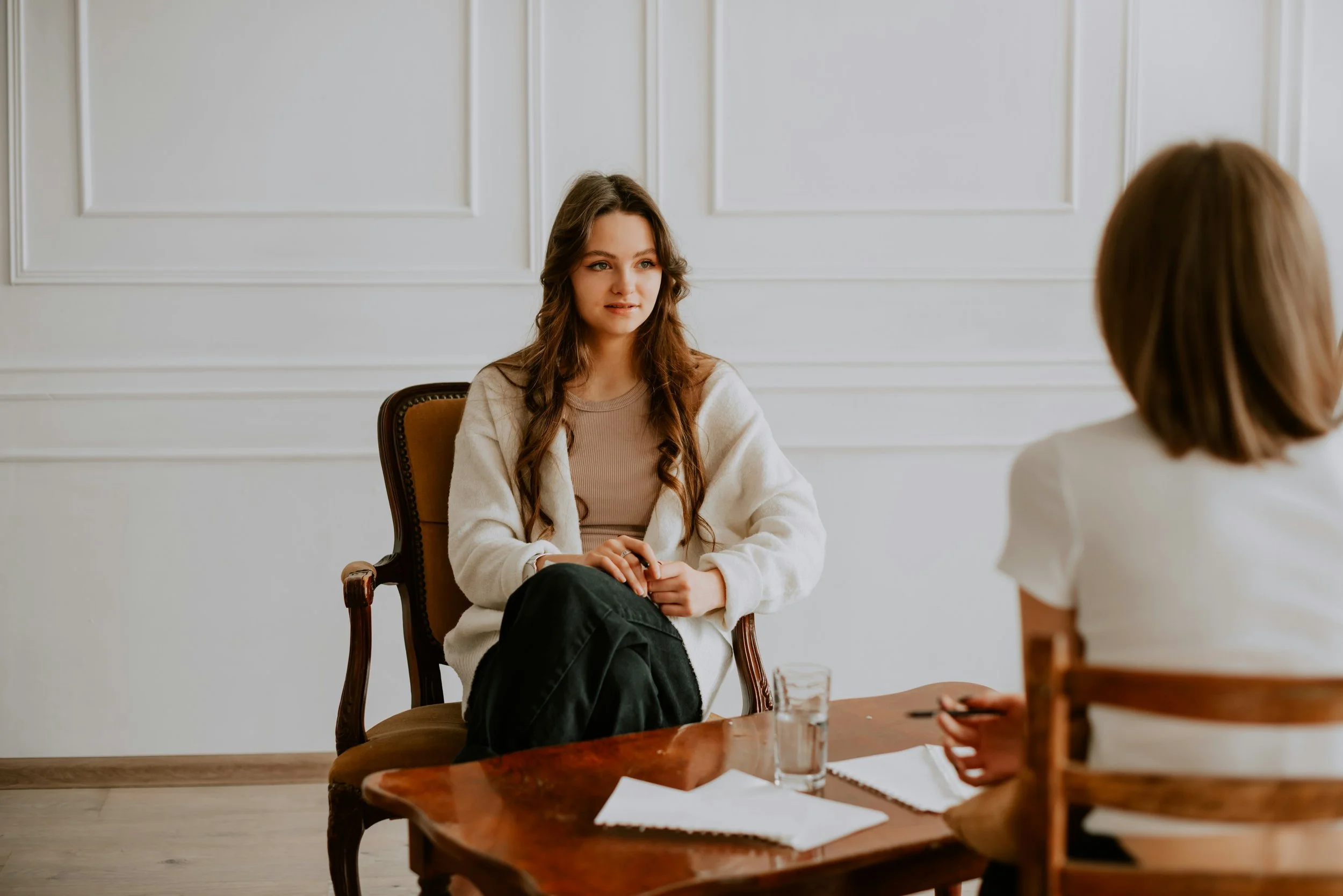 A young woman with long, wavy brown hair sitting on a chair at a table, engaged in conversation with another woman who has short, blonde hair, in a bright, white room with paneled walls.