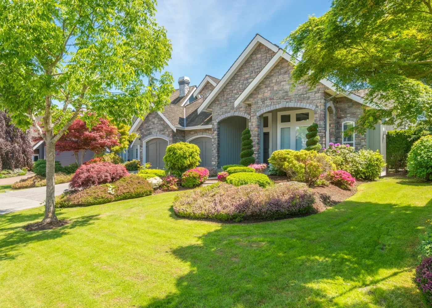 A well-maintained front yard with lush green grass, colorful flowering bushes, and trees in front of a stone house with arched garage doors and a gabled roof.