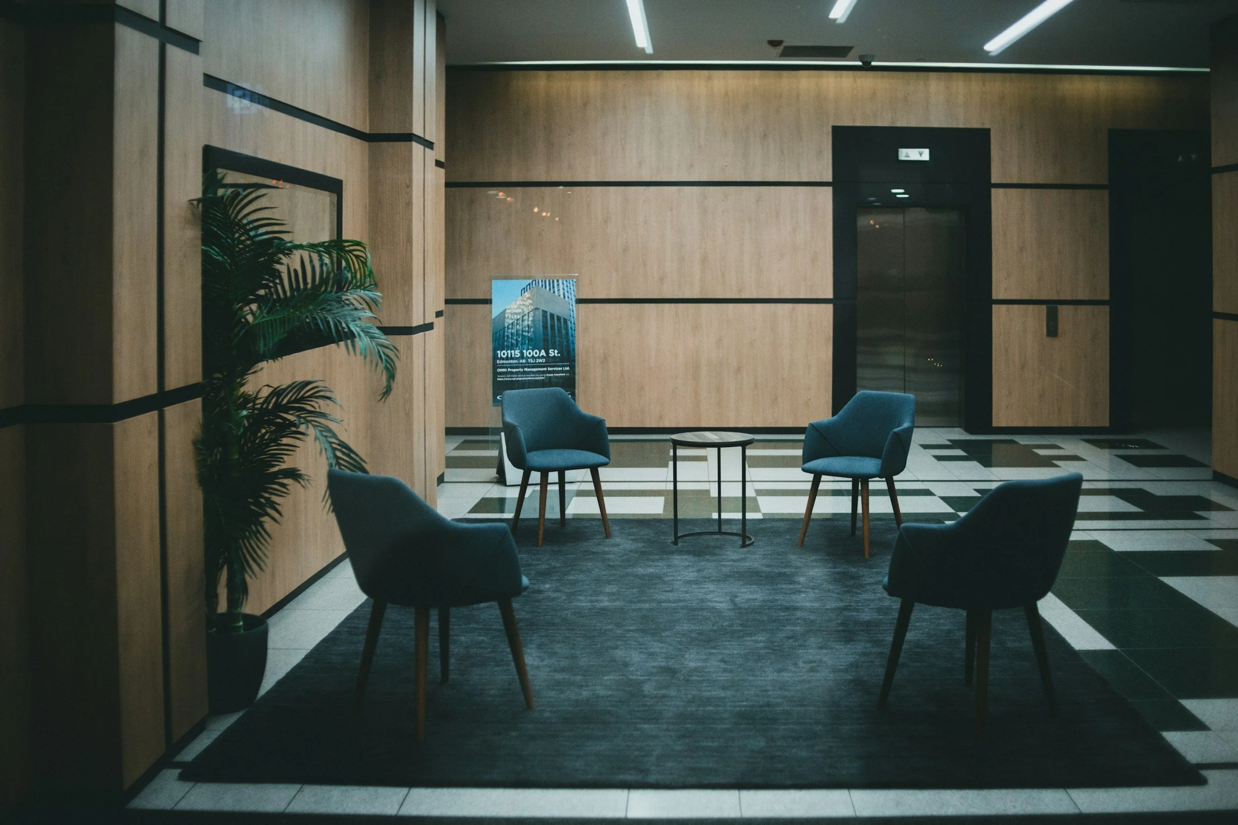 A modern lobby with wood-paneled walls, four blue armchairs arranged around a small round table on a dark rug, a potted plant on the left, and an elevator in the background.