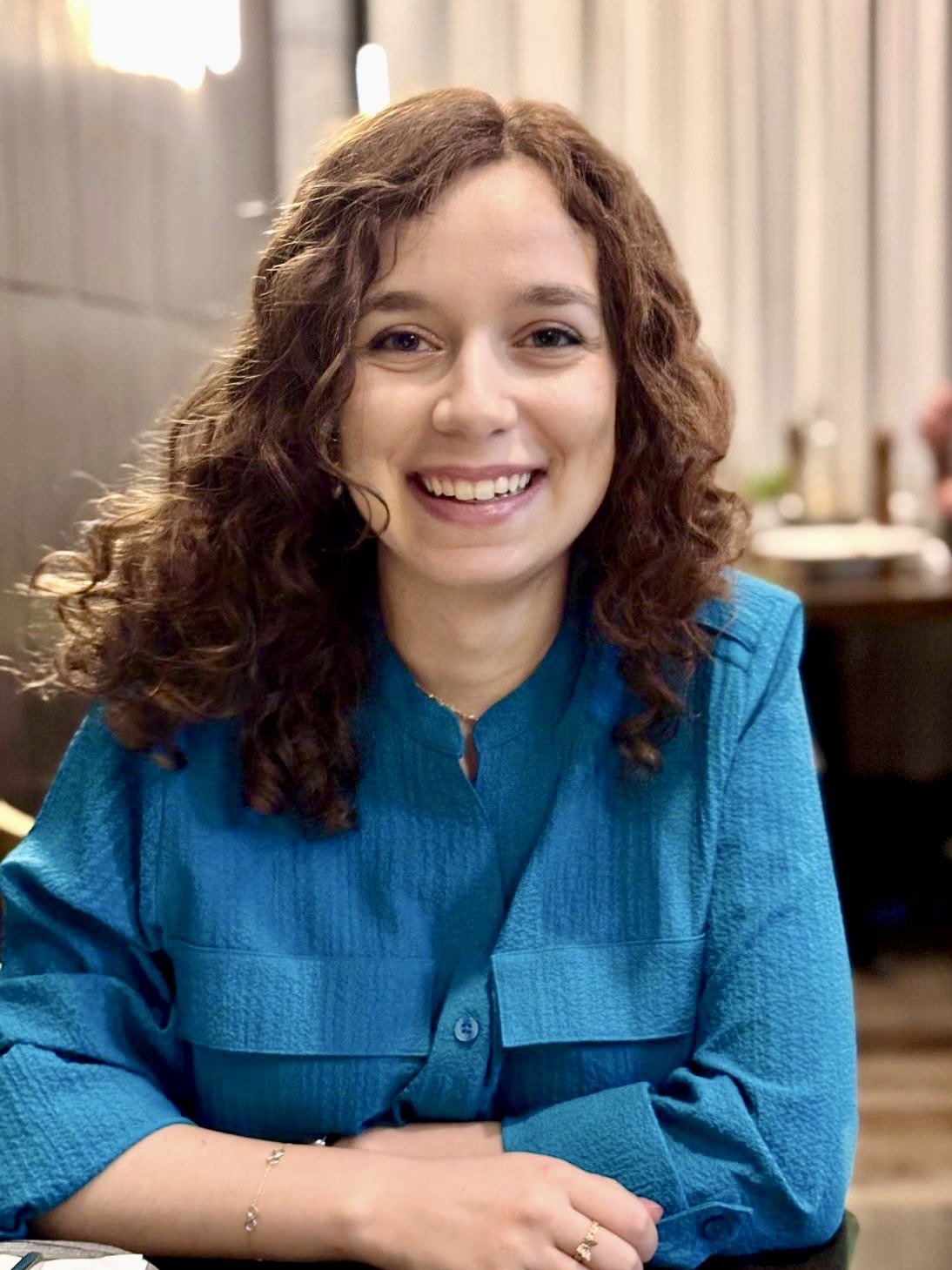 A woman with curly brown hair smiling, wearing a blue blouse, sitting indoors.