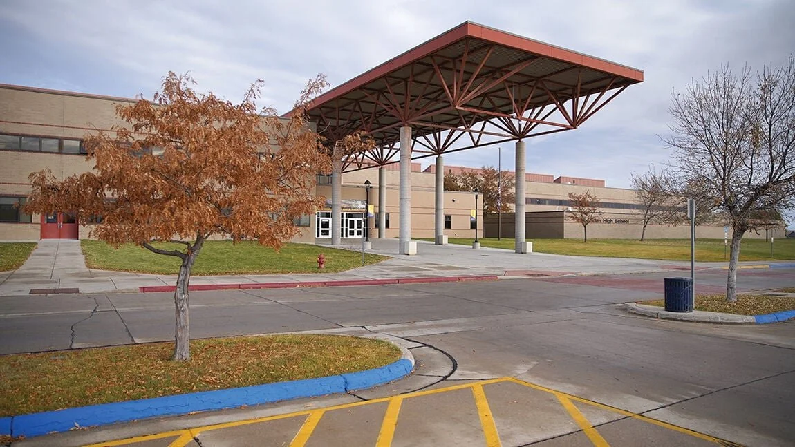 Empty school parking lot with trees, sidewalk, and a large building with a distinctive red roofed canopy.