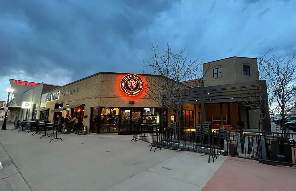 Exterior view of a restaurant with a neon sign, outdoor seating, and leafless trees, evening sky in the background.