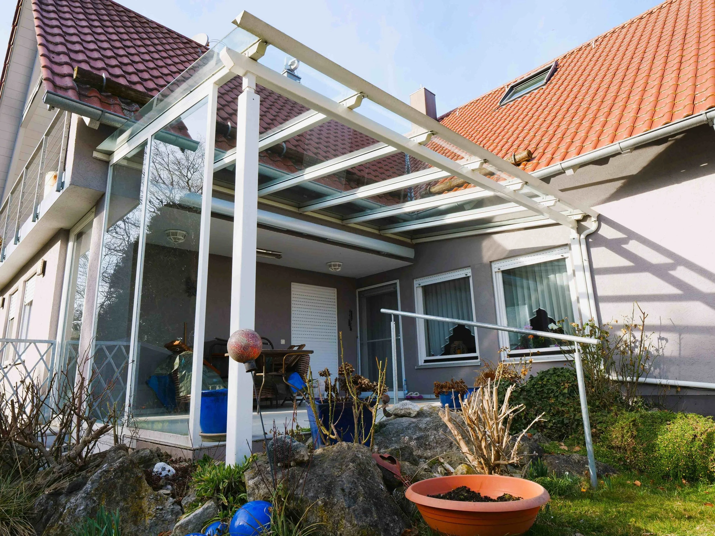 Garten mit Steinen, Pflanzen, Blumentopf und einem verglasten Wintergarten an einem Haus mit roten Dachziegeln.