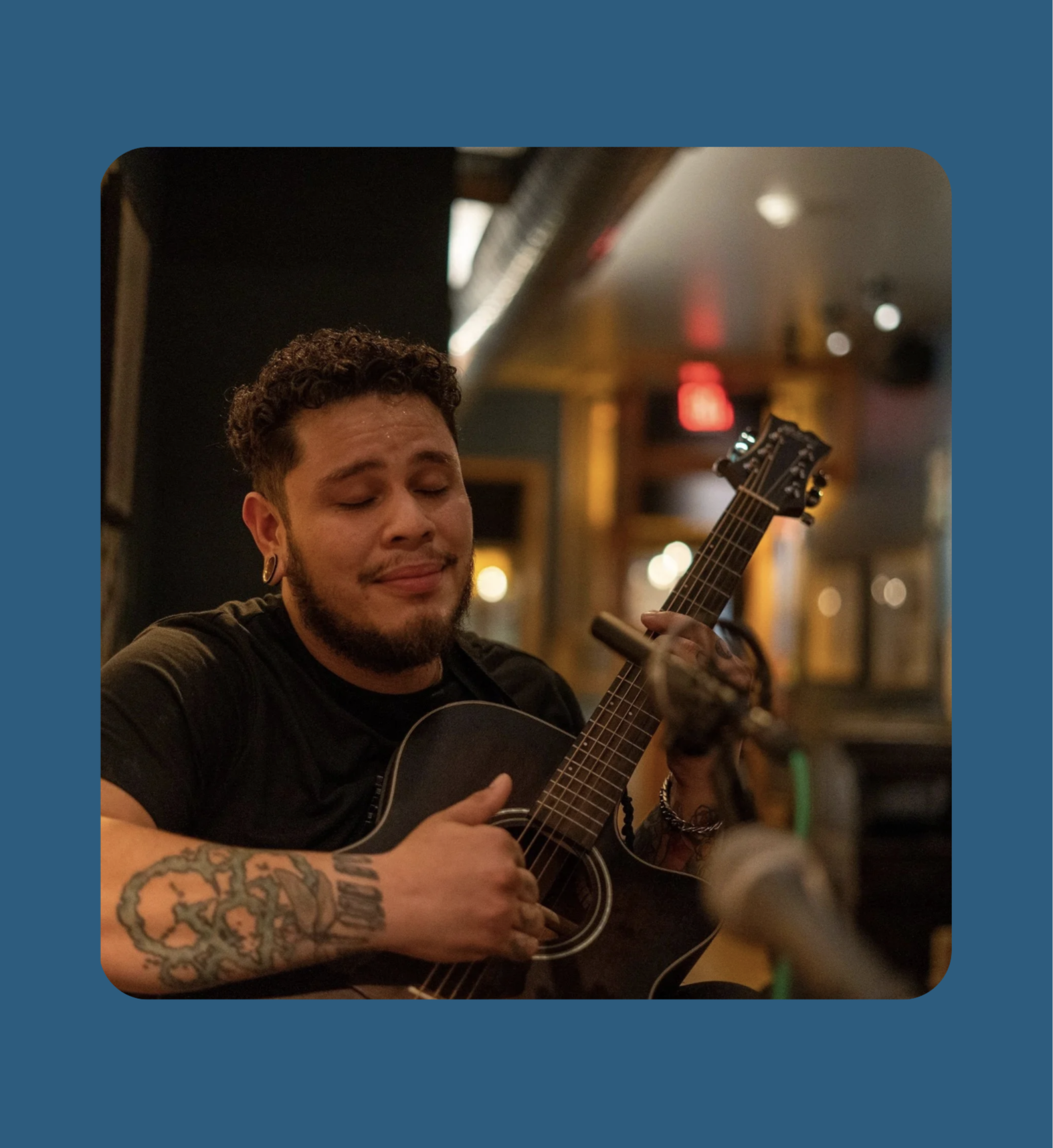 A man with tattoos and earrings playing an acoustic guitar in an intimate venue near Fishtown.