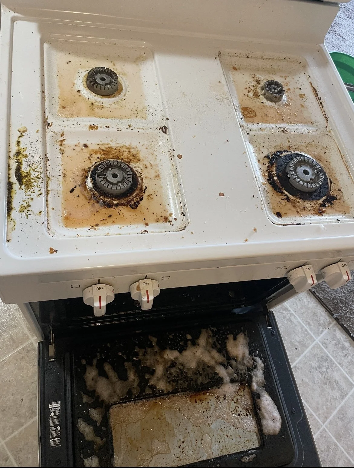 A dirty four-burner stove with burnt food and grease, with an oven beneath filled with burnt and melted food residue and grease stains.