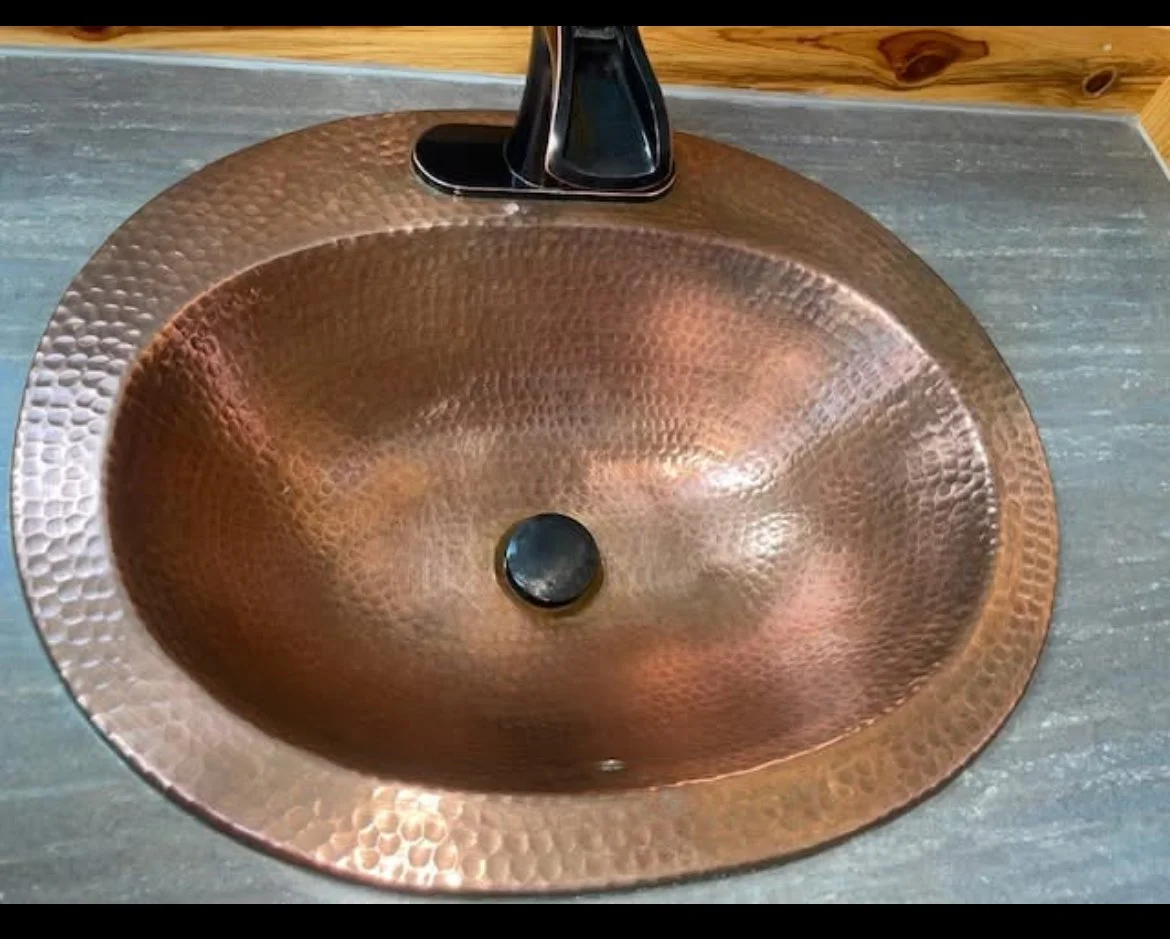 Close-up of a metallic copper-colored oval bathroom sink with a hammered texture, mounted on a gray countertop with a modern black faucet attached.