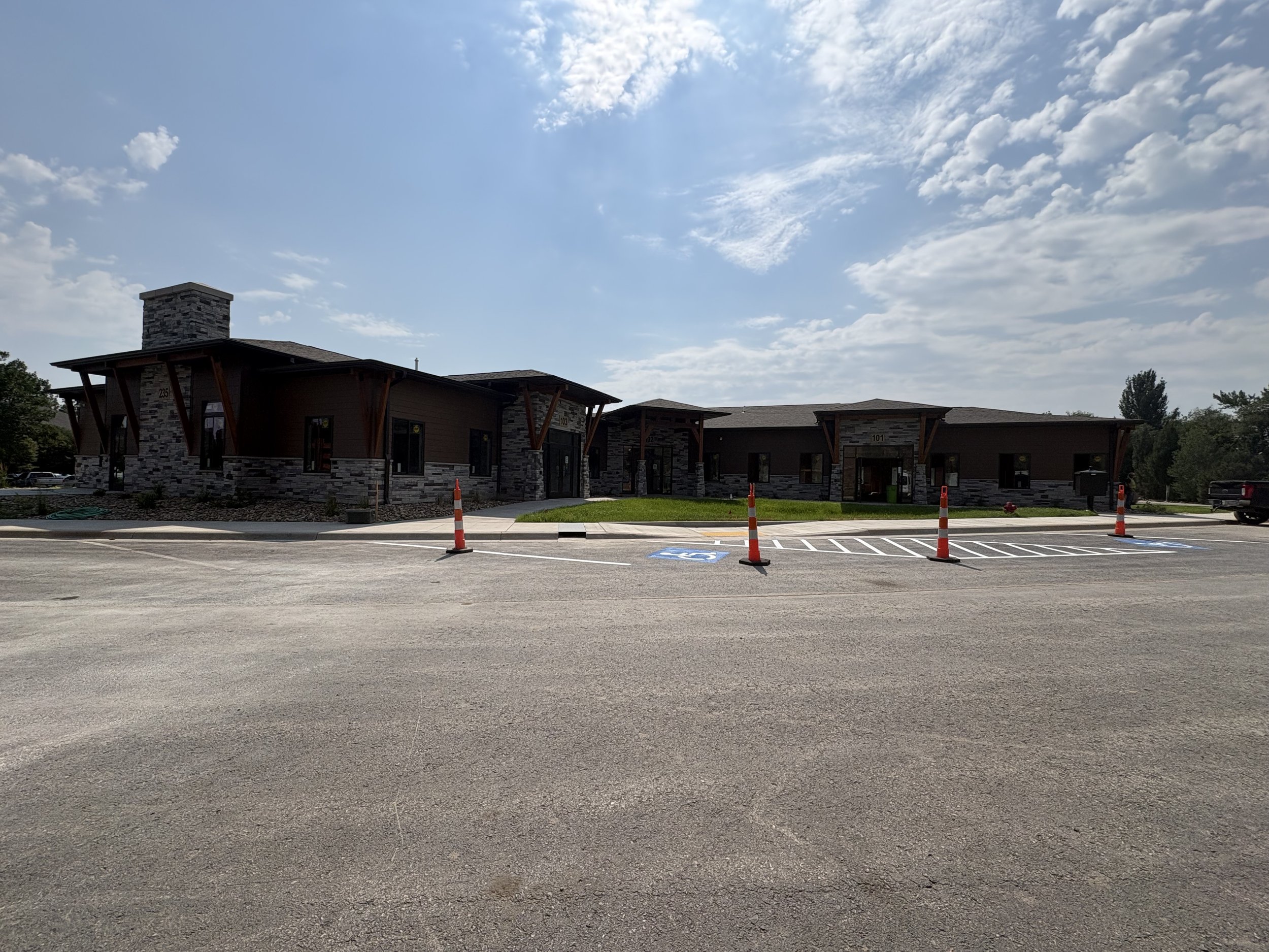 Image of a modern building with stone accents and a parking lot in front. The parking lot has reserved and accessible parking spaces marked by painted signs and orange cones. The sky is partly cloudy with white clouds scattered across a blue sky.