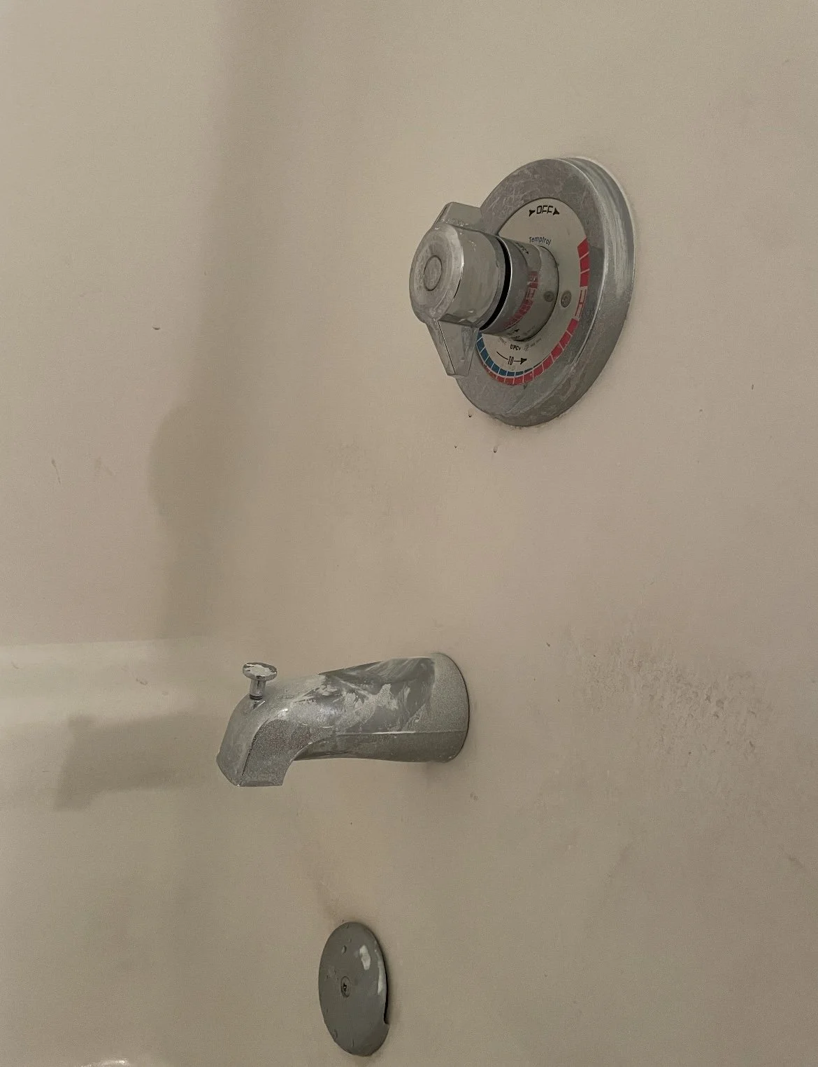 A shower with a metal control knob, a valve, and a drain on the tub floor. The wall and fixtures are somewhat aged and stained.