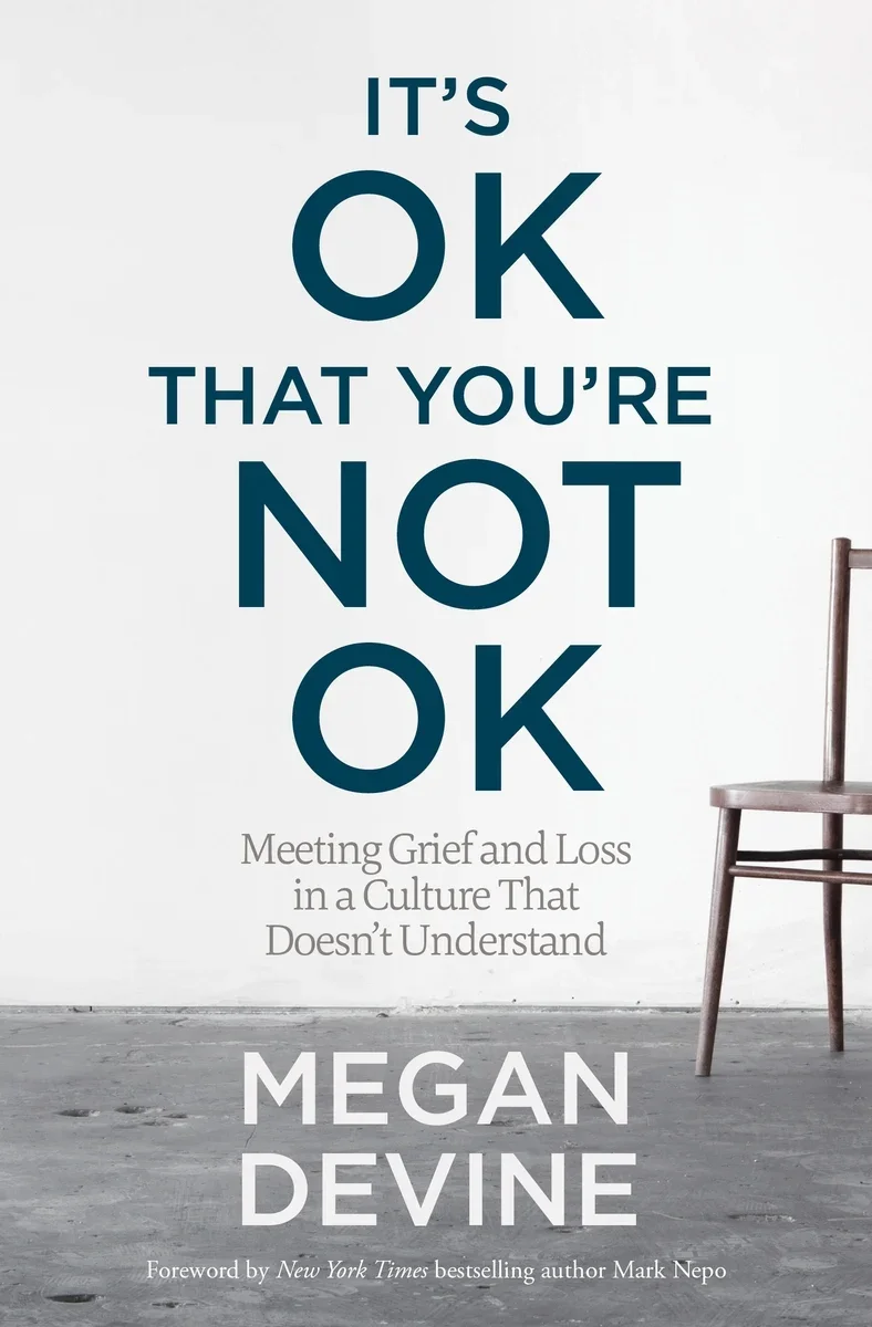 Book cover titled 'It's OK That You're Not OK' by Megan Devine, with subtitle 'Meeting Grief and Loss in a Culture That Doesn't Understand,' and a foreword by Mark Nepo. The cover features a simple image of a wooden chair against a white wall and a gray concrete floor.