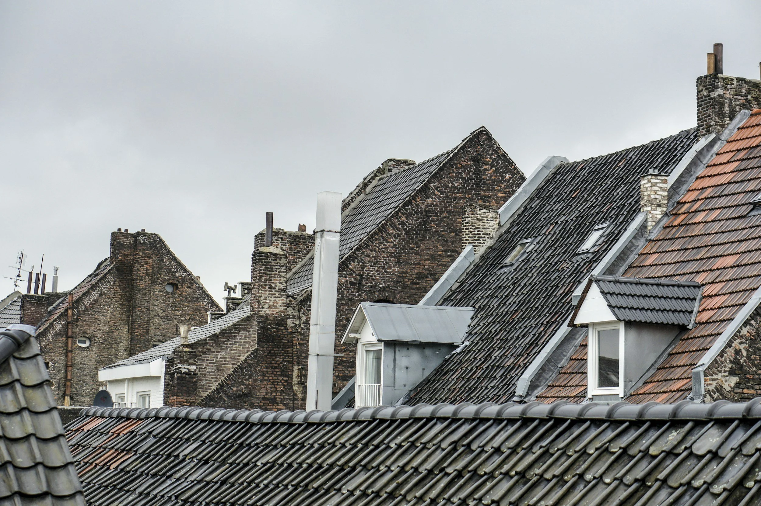 Close-up view of rooftops with variously aged and styled tiles and bricks under cloudy sky.