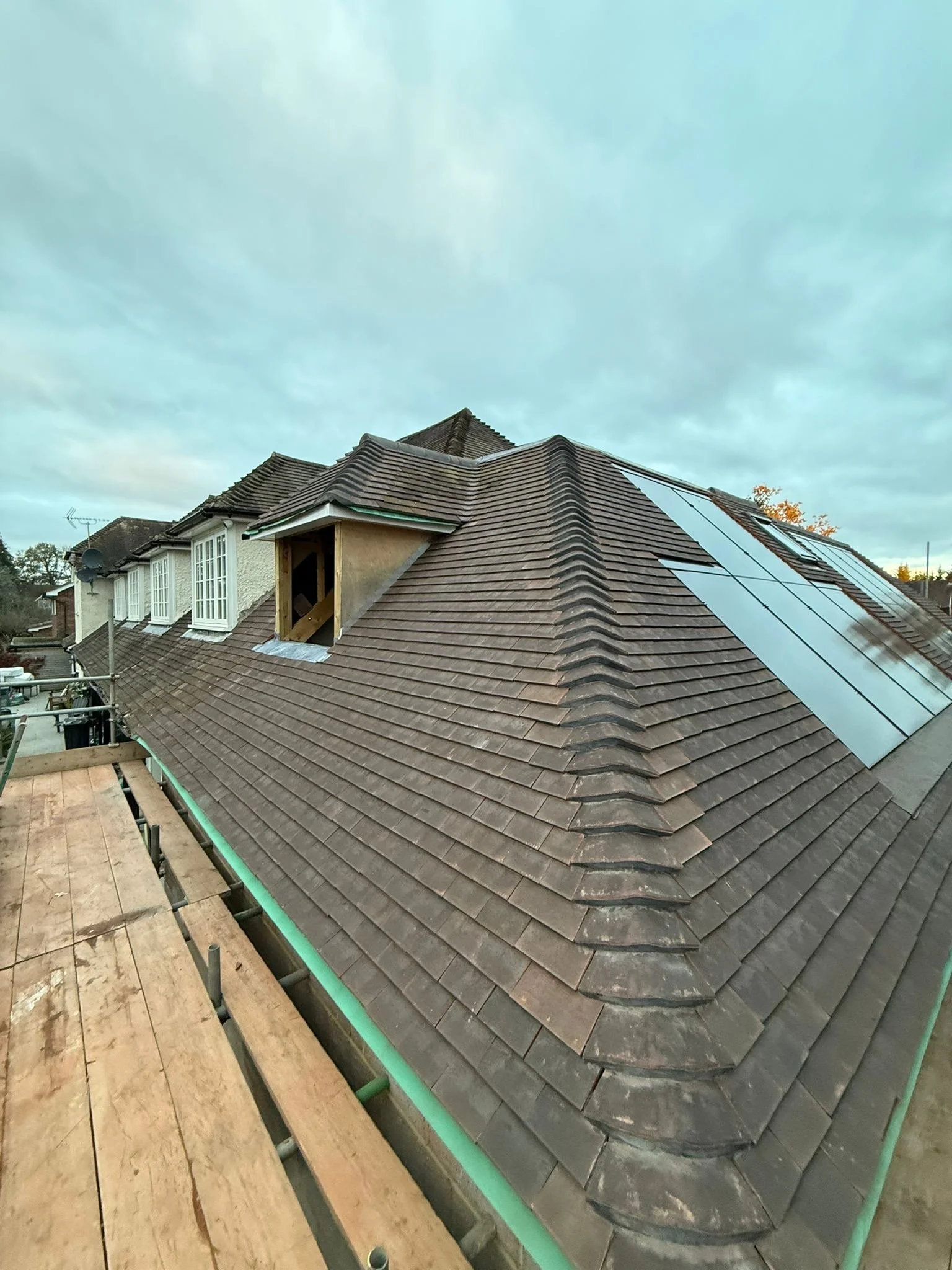 View of residential roof with brown tiles, some new solar panels, and a small dormer window under construction, with scaffolding and partly cloudy sky in the background.