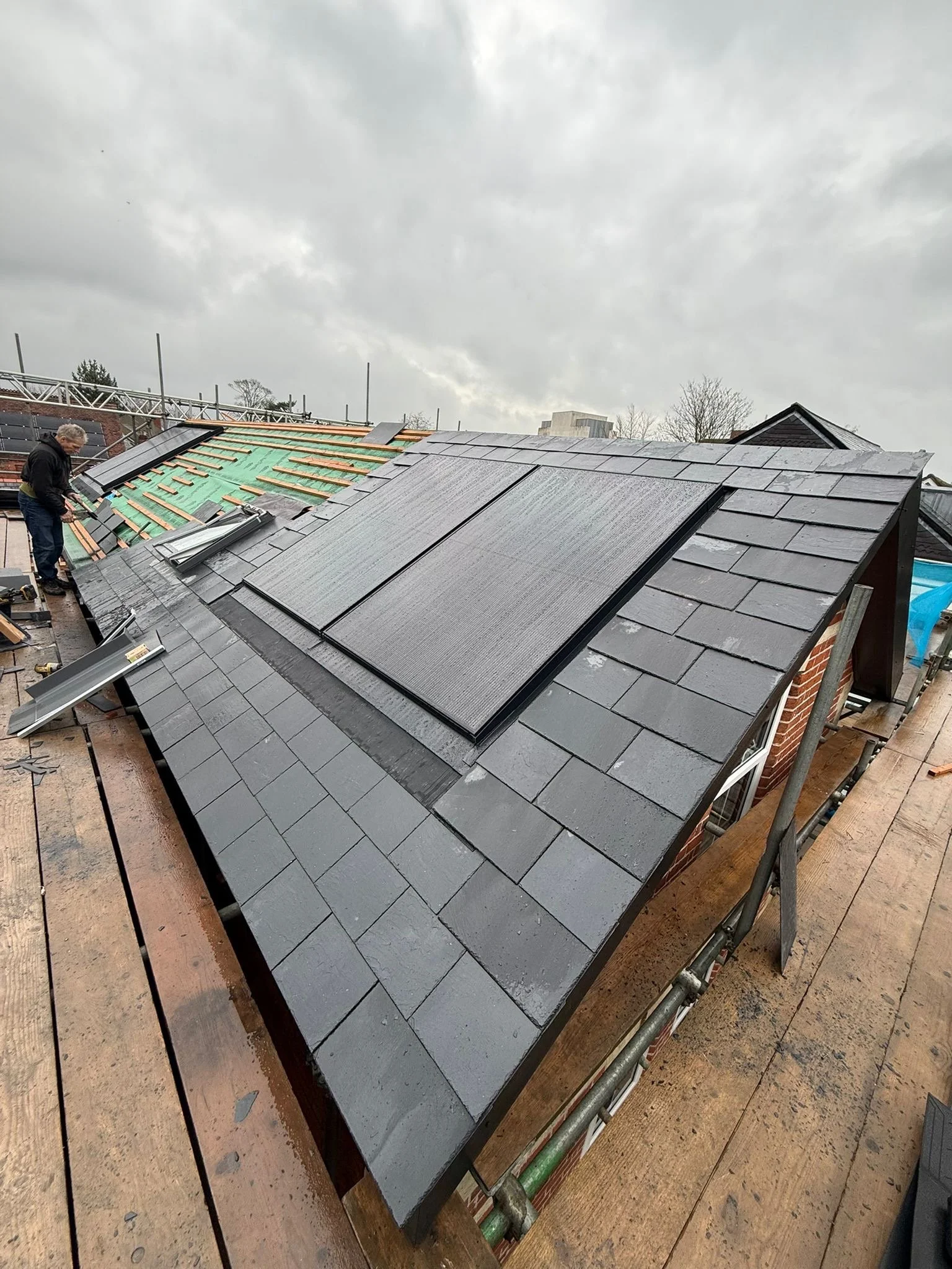 A construction worker installing black roof tiles on a sloped roof. Part of the roof is still under construction, with green underlayment visible. The sky is cloudy and overcast.