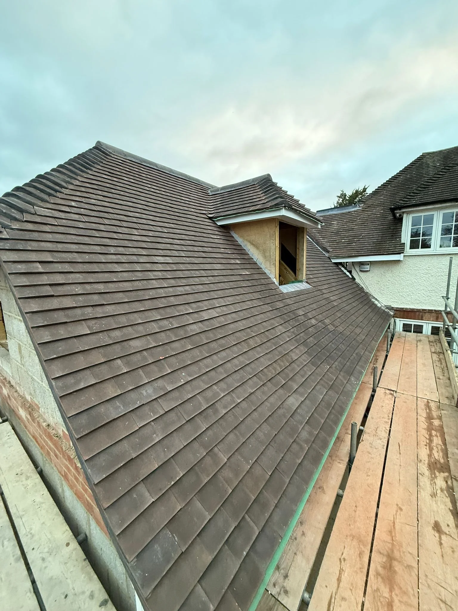 Newly installed brown roof shingles on a house with exterior framing and a small dormer opening, surrounded by scaffolding and a wooden walkway.