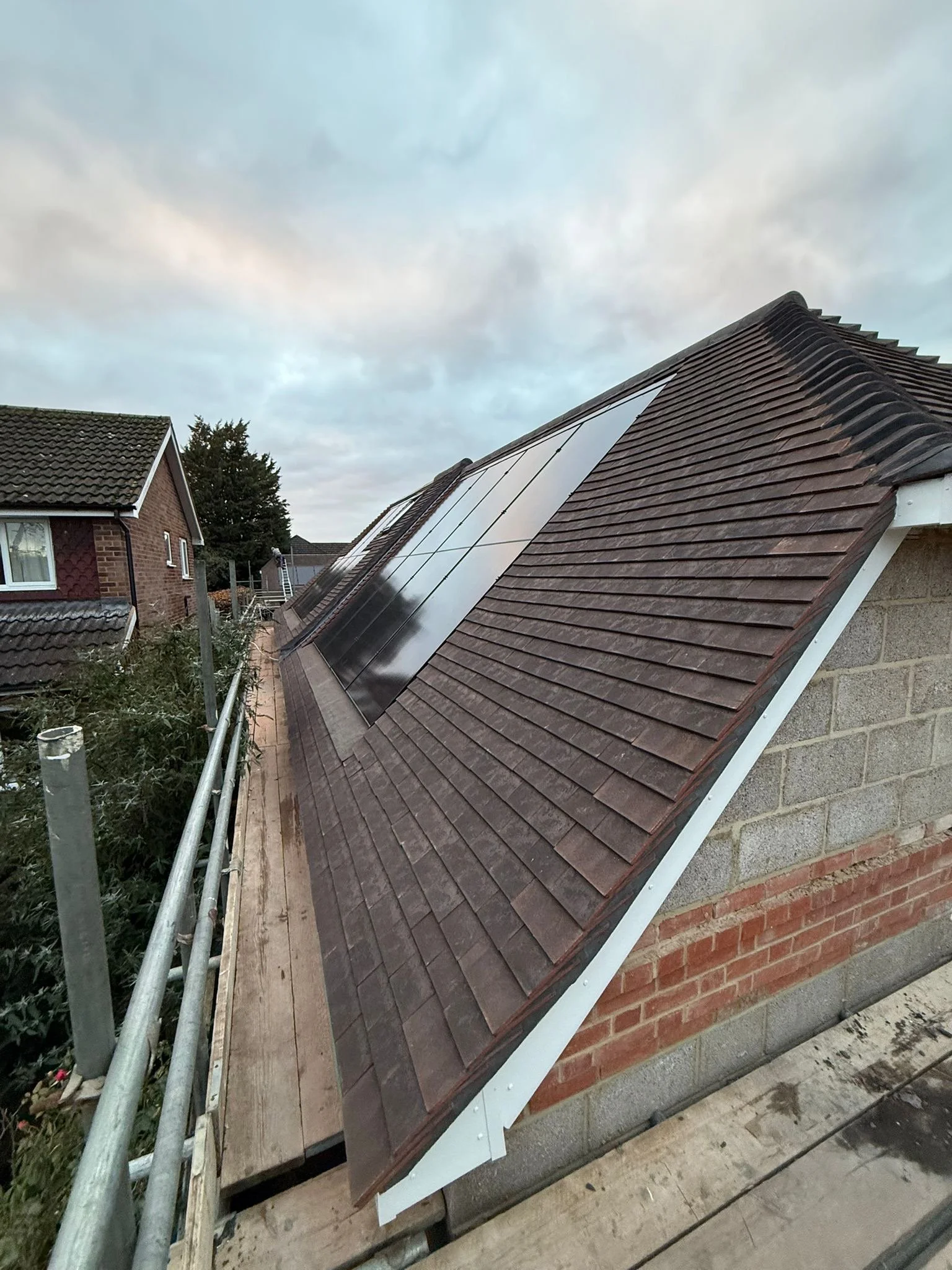 A rooftop with solar panels installed on a sloped roof of a house, with neighboring houses and trees in the background under a cloudy sky.