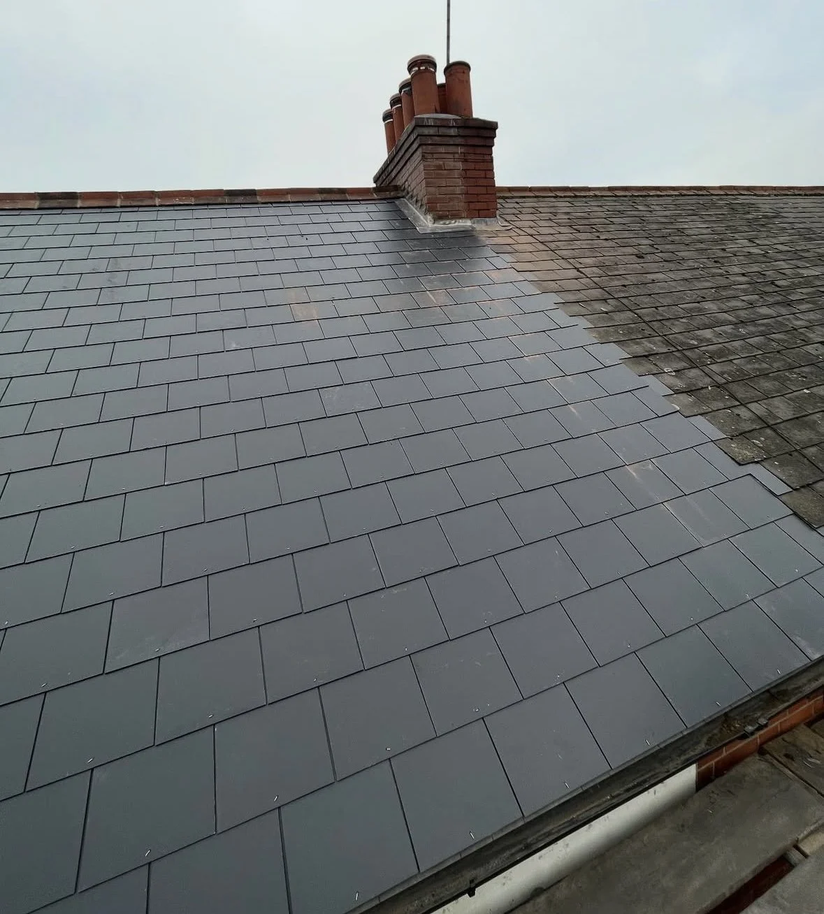 A rooftop showing a restoration process with new dark gray tiles replacing old weathered tiles, and a chimney with four red clay chimney pots.