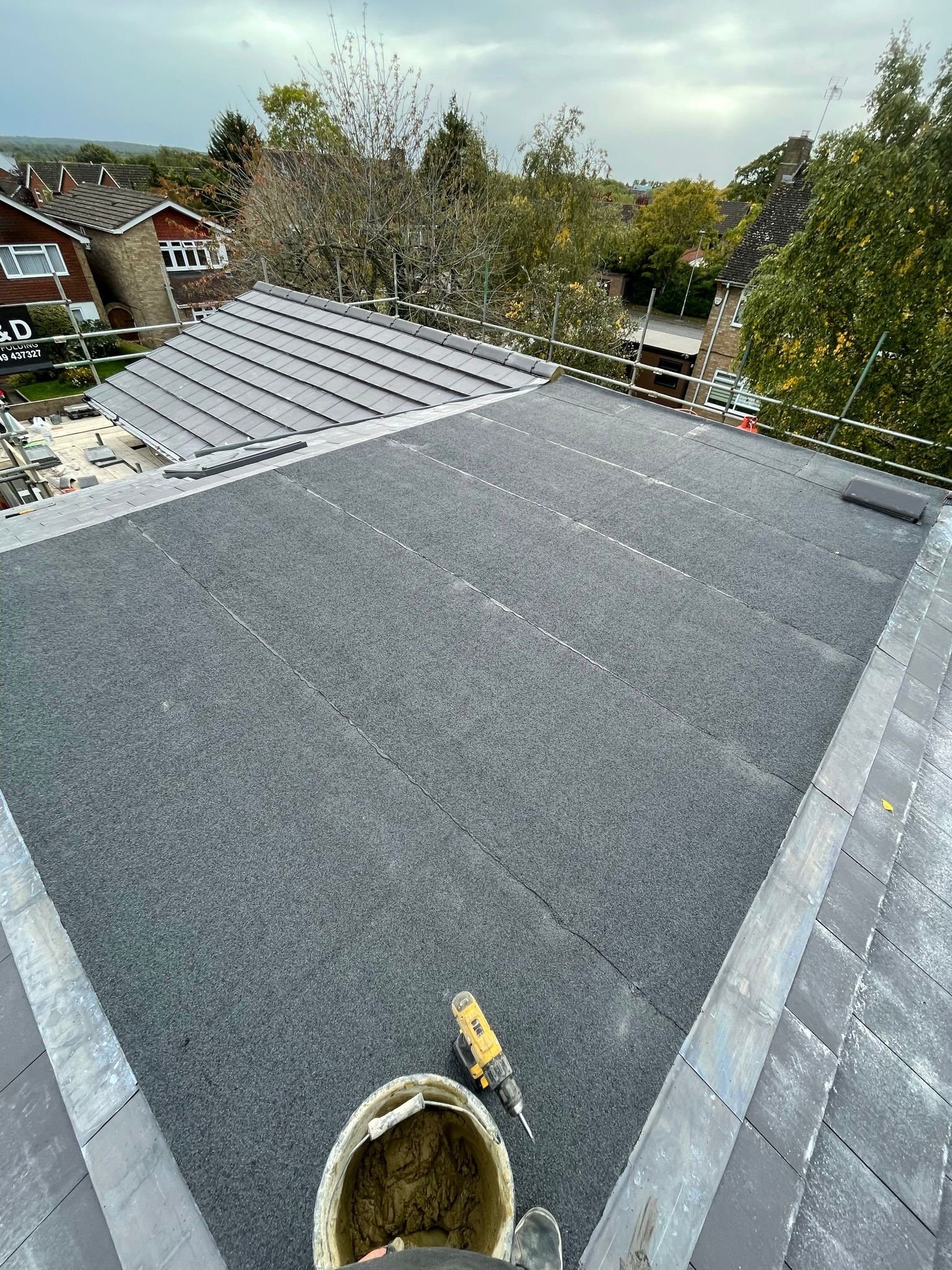 View from a rooftop under construction with a new layer of roofing felt, surrounding scaffolding, and neighboring houses and trees seen in a suburban neighborhood.