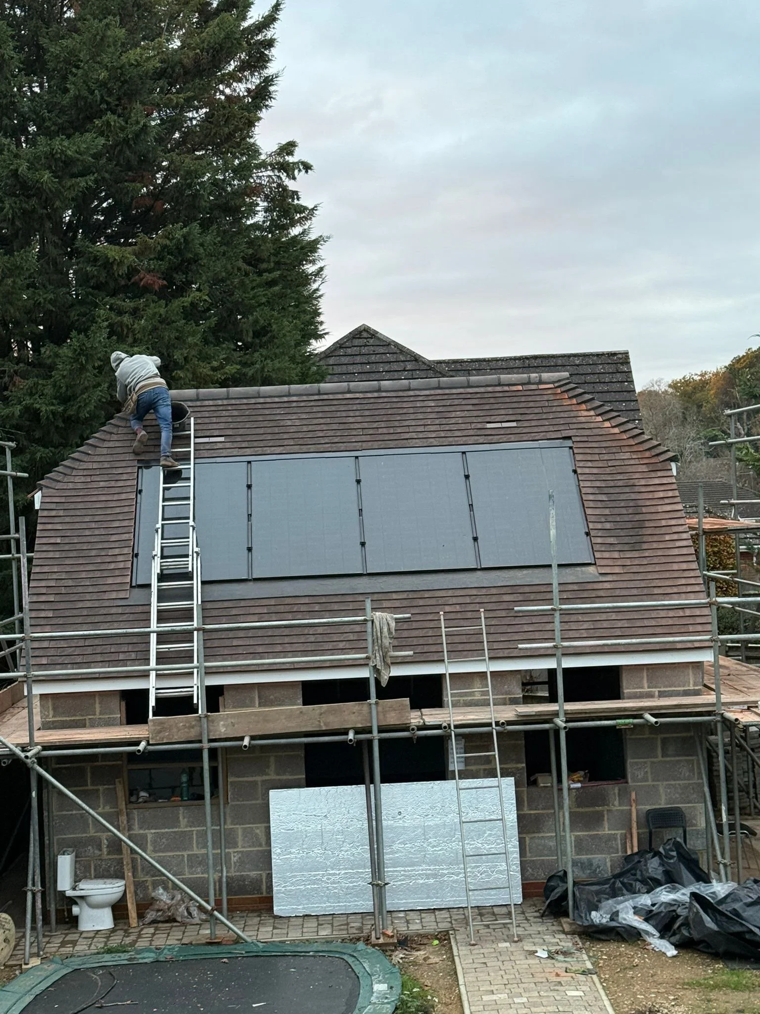 A person working on installing solar panels on the roof of a house under construction, with scaffolding surrounding the building, and a small trampoline in the yard. CPS ROOFING IMAGE