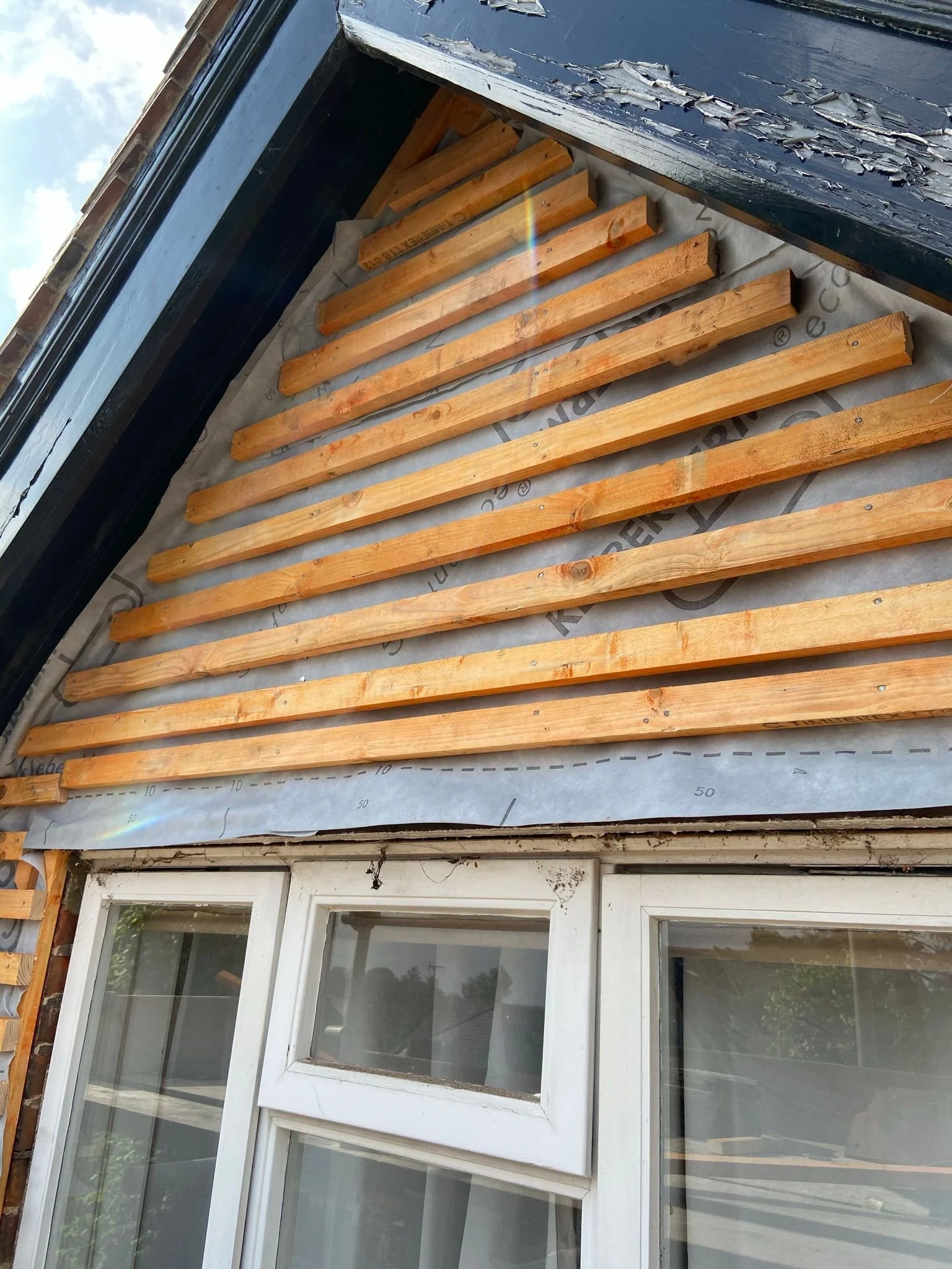 Construction of a house exterior with mini horizontal wooden slats installed above a window, and peeling paint on the upper wall.