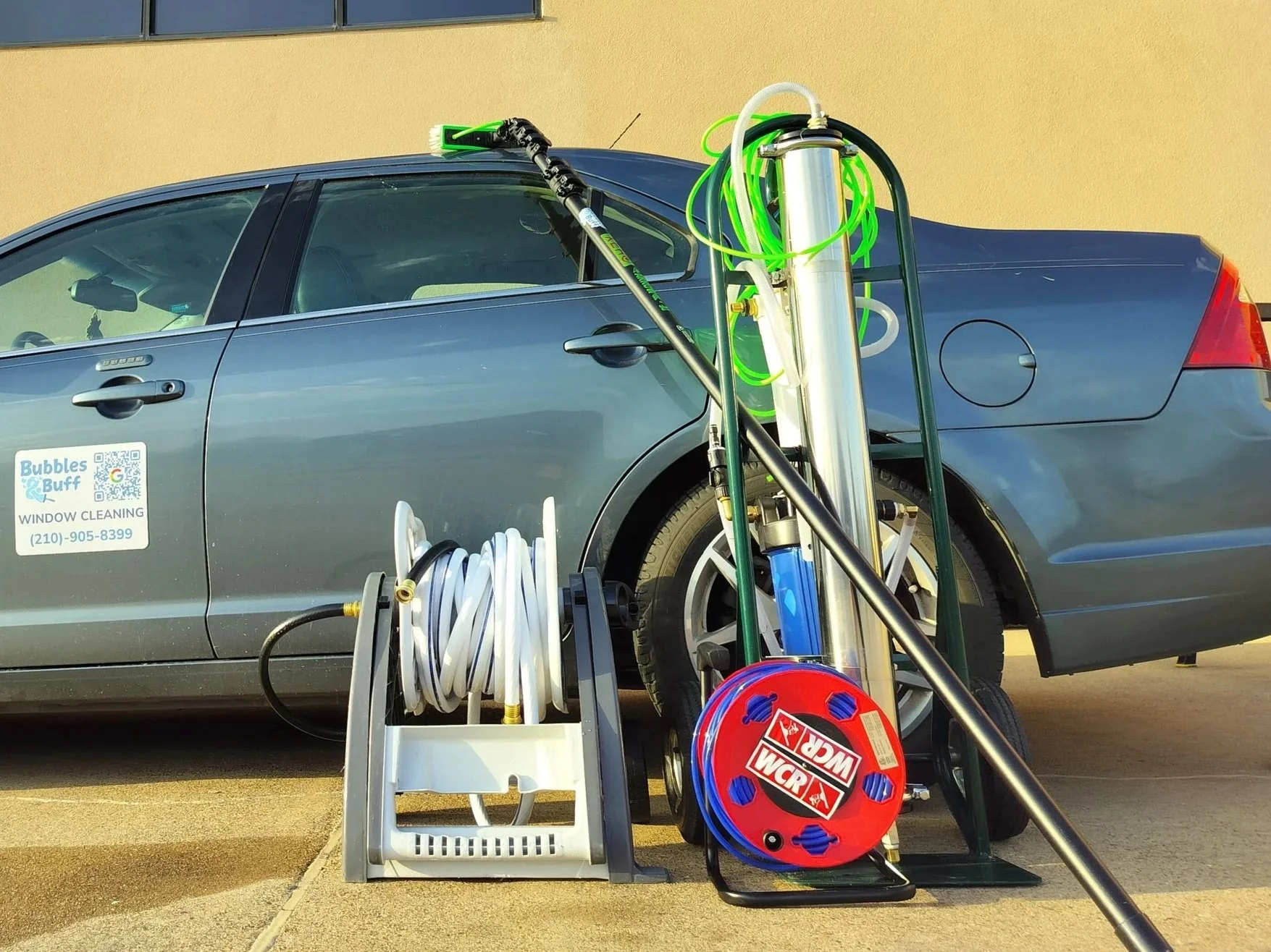 A window cleaning service vehicle parked on a concrete surface with equipment for high-rise window cleaning, including hoses, a pole, and a vacuum or pump system, positioned next to the car.