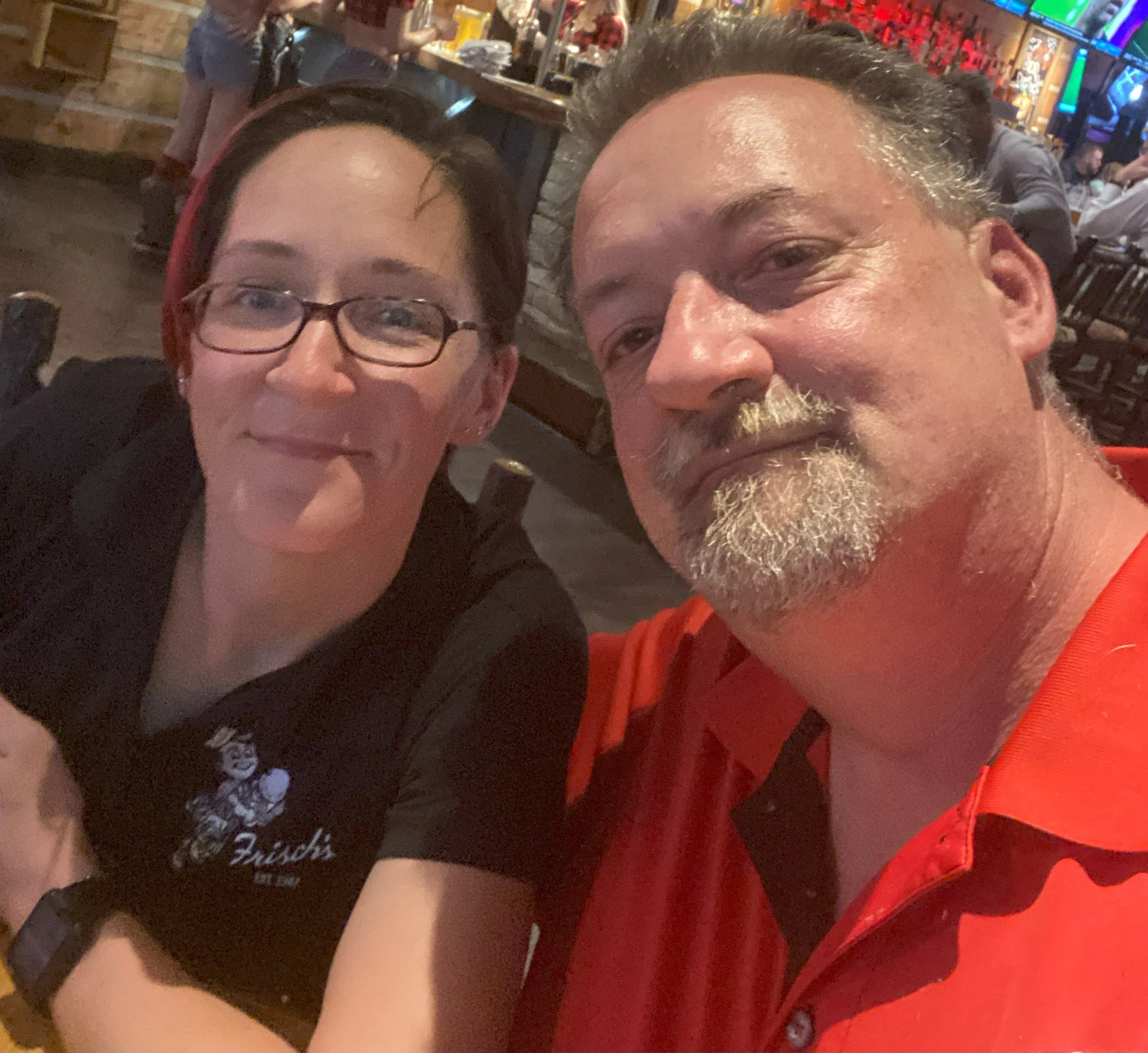 A man and a woman taking a selfie in a restaurant or bar. The woman has glasses and dark hair tied back, the man has a beard and short hair. The background shows a lively indoor setting with other people and bright lights.