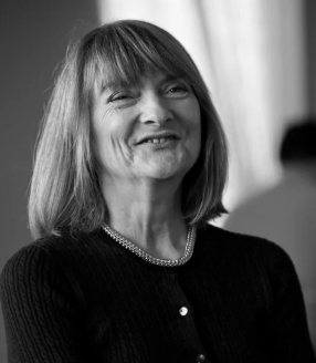 Black and white photo of a smiling woman with shoulder-length hair, wearing a dark top and a necklace, indoors near a window.