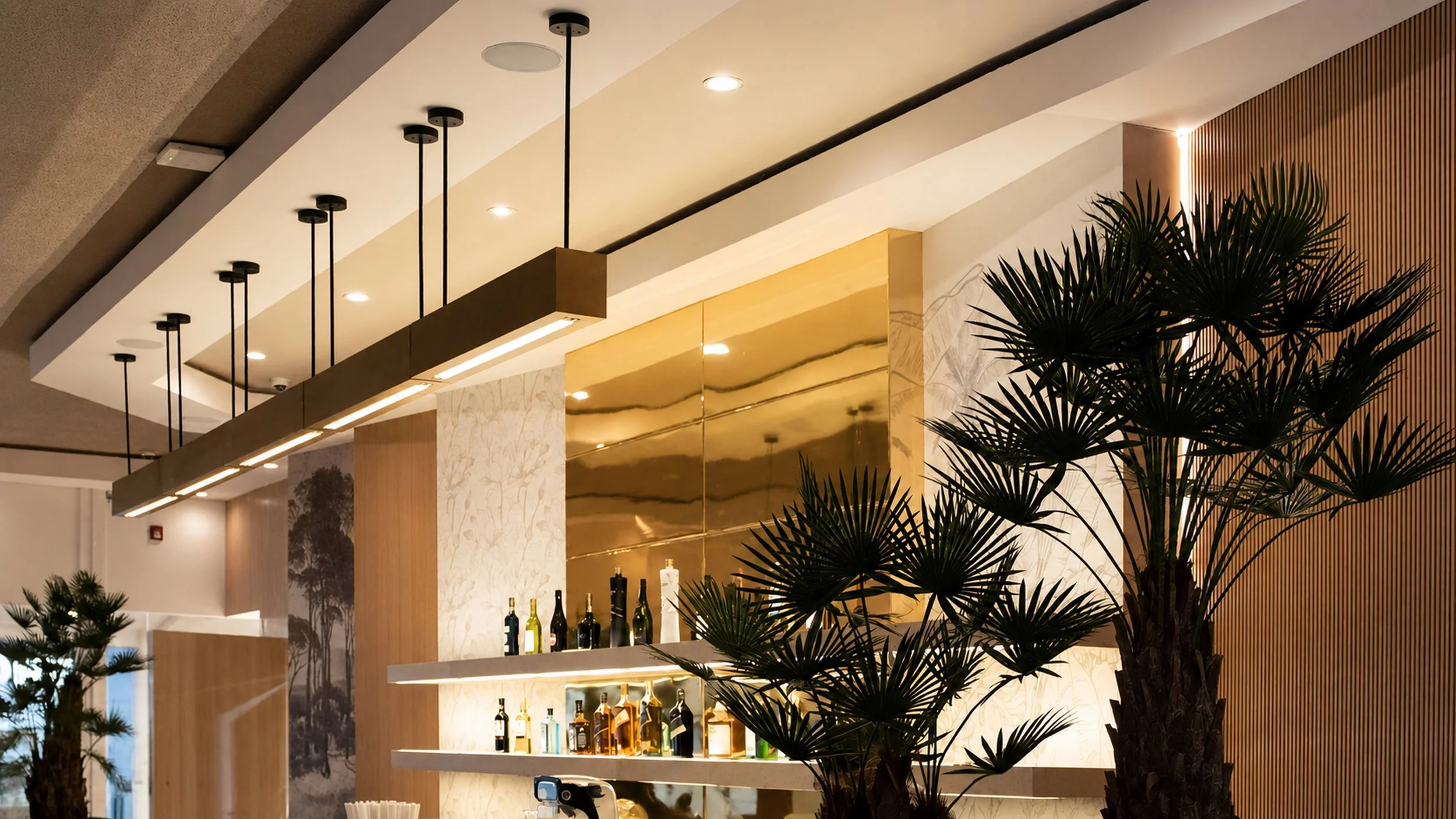 Interior de un espacio con decoración moderna, plantas grandes, área de bar con botellas y una iluminación suspendida.