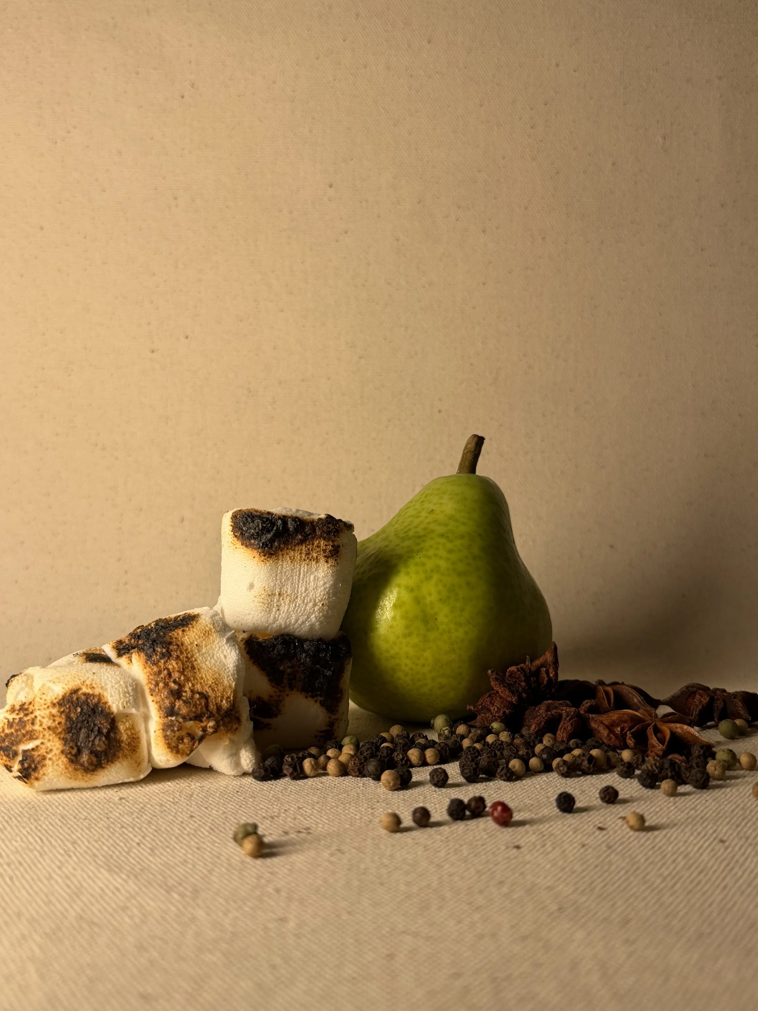 A green pear, toasted marshmallows, star anise, and mixed peppercorns arranged on a beige surface.