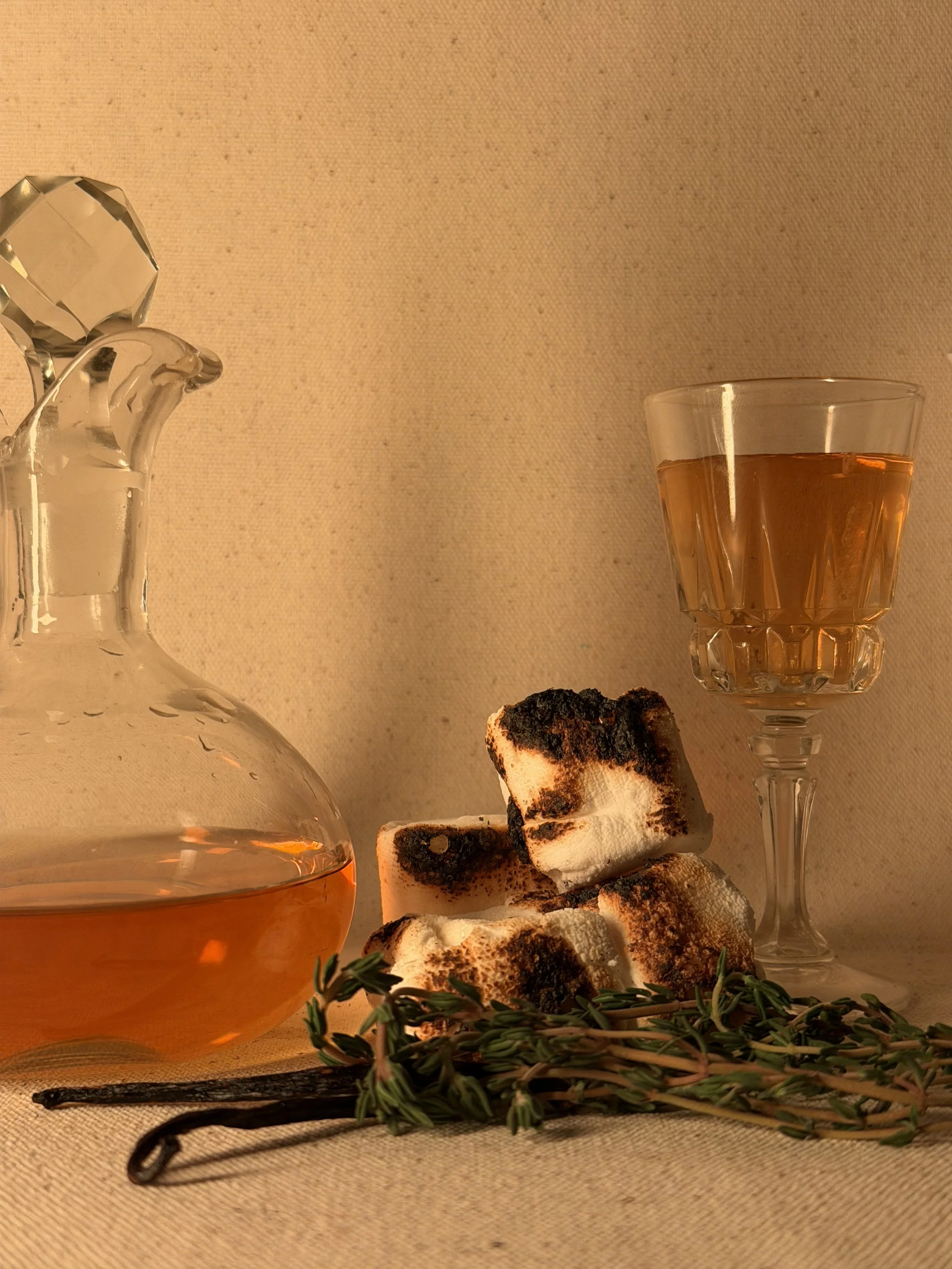 A crystal decanter with a honey-colored liquid, a glass of similar liquid on a tall stemmed glass, toasted marshmallows with burnt tops, fresh thyme sprigs, and a vanilla bean on a beige surface with a beige background.