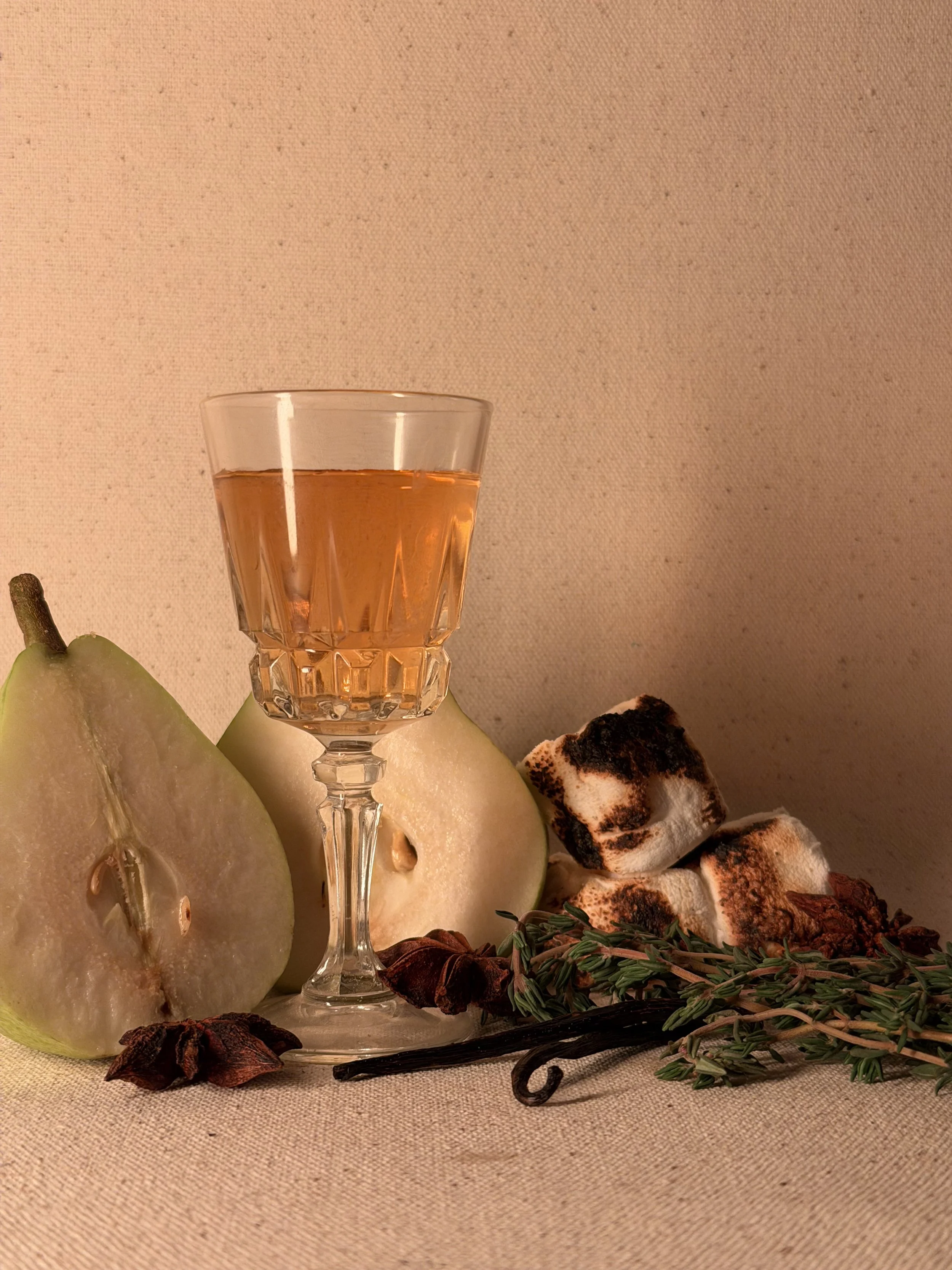 A glass of amber-colored liquid on a table with sliced pears, toasted marshmallows, a vanilla bean, and sprigs of thyme, set against a beige background.