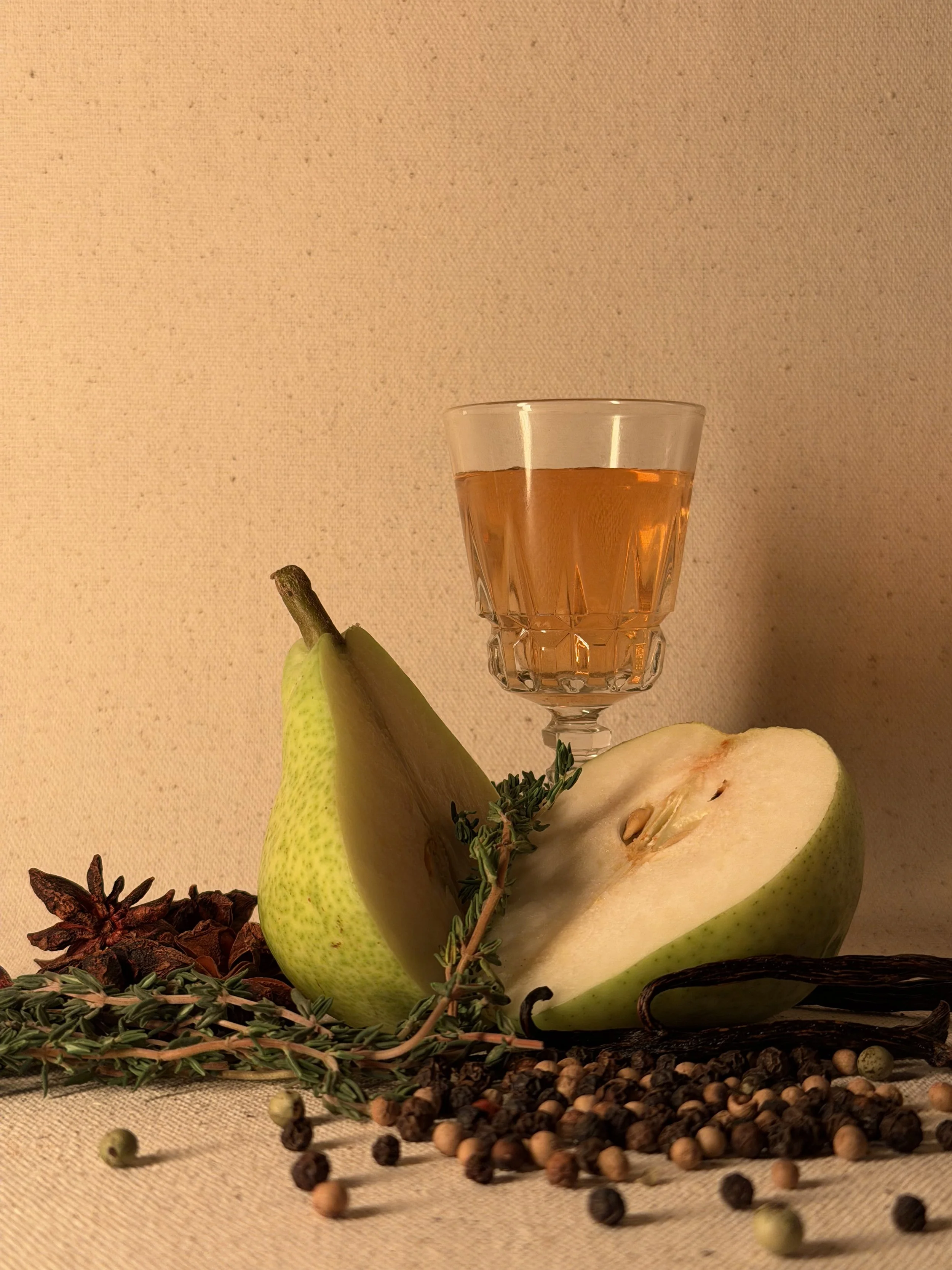 Half-cut green pear, a glass of amber liquid, dried herbs, peppercorns, and spices scattered on a beige surface.