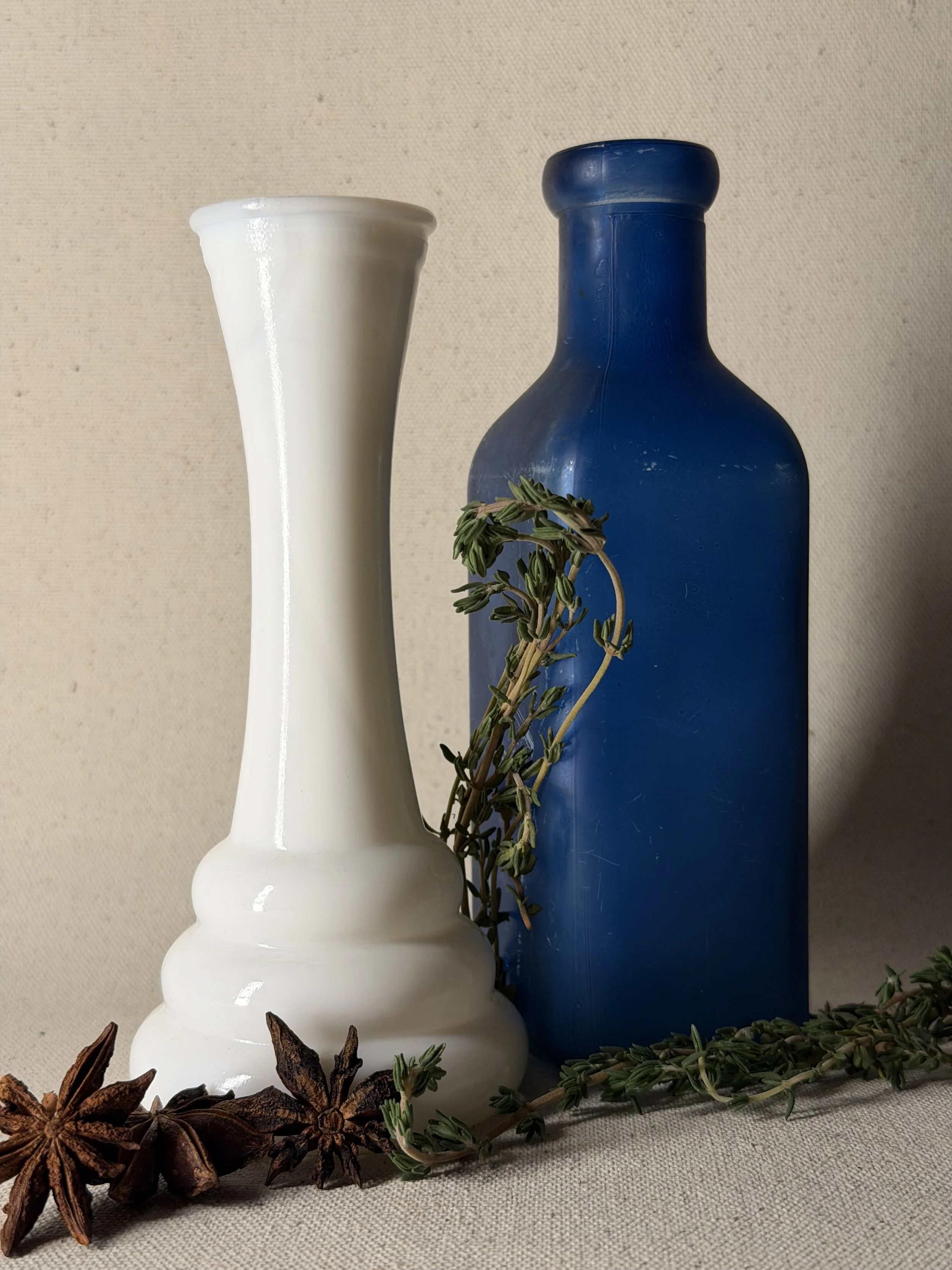 A white ceramic vase and a dark blue bottle are displayed against a neutral background, with sprigs of green thyme and star anise placed near the base.