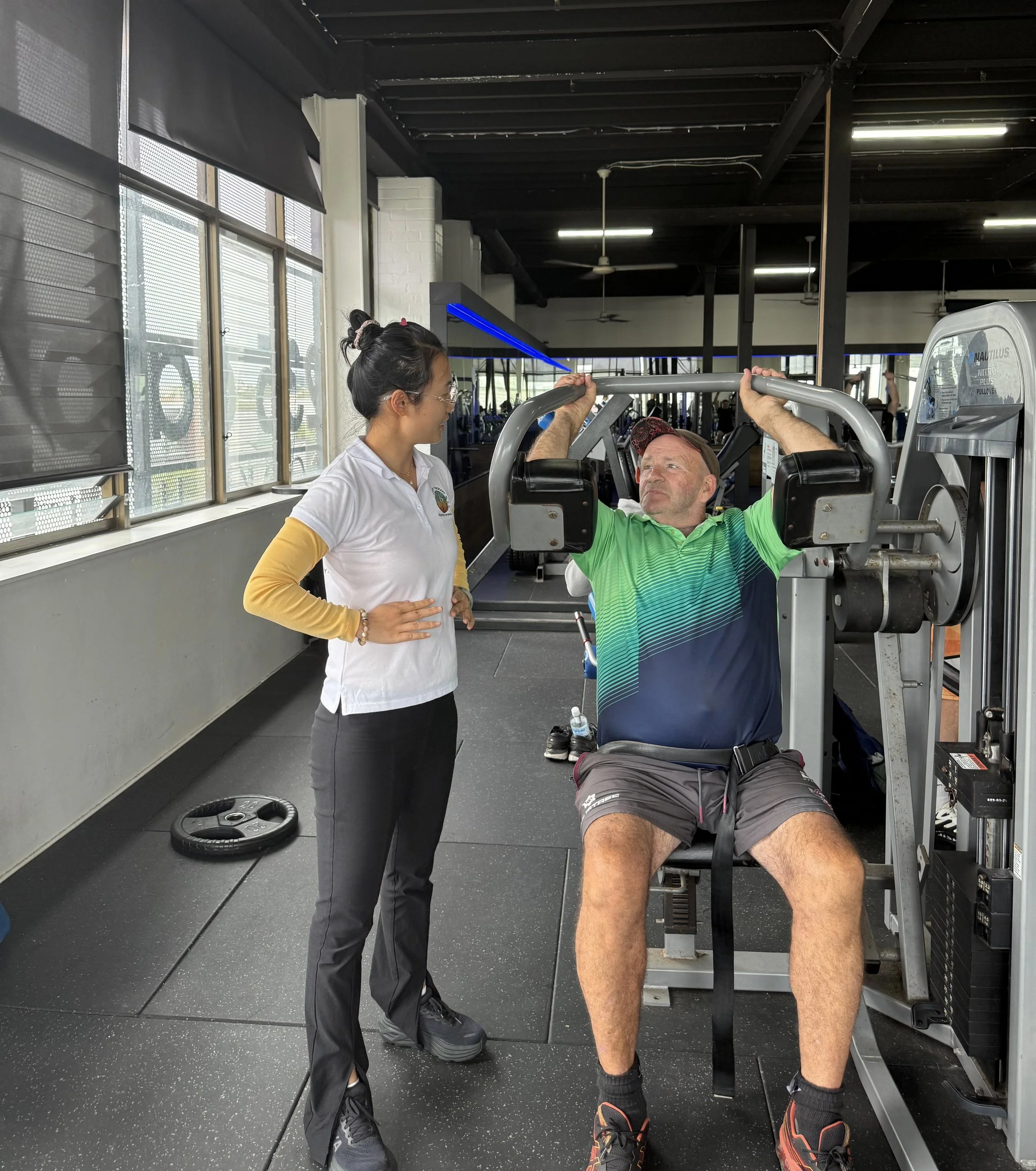 A woman trainer assisting a man with a seated shoulder press exercise at the gym.