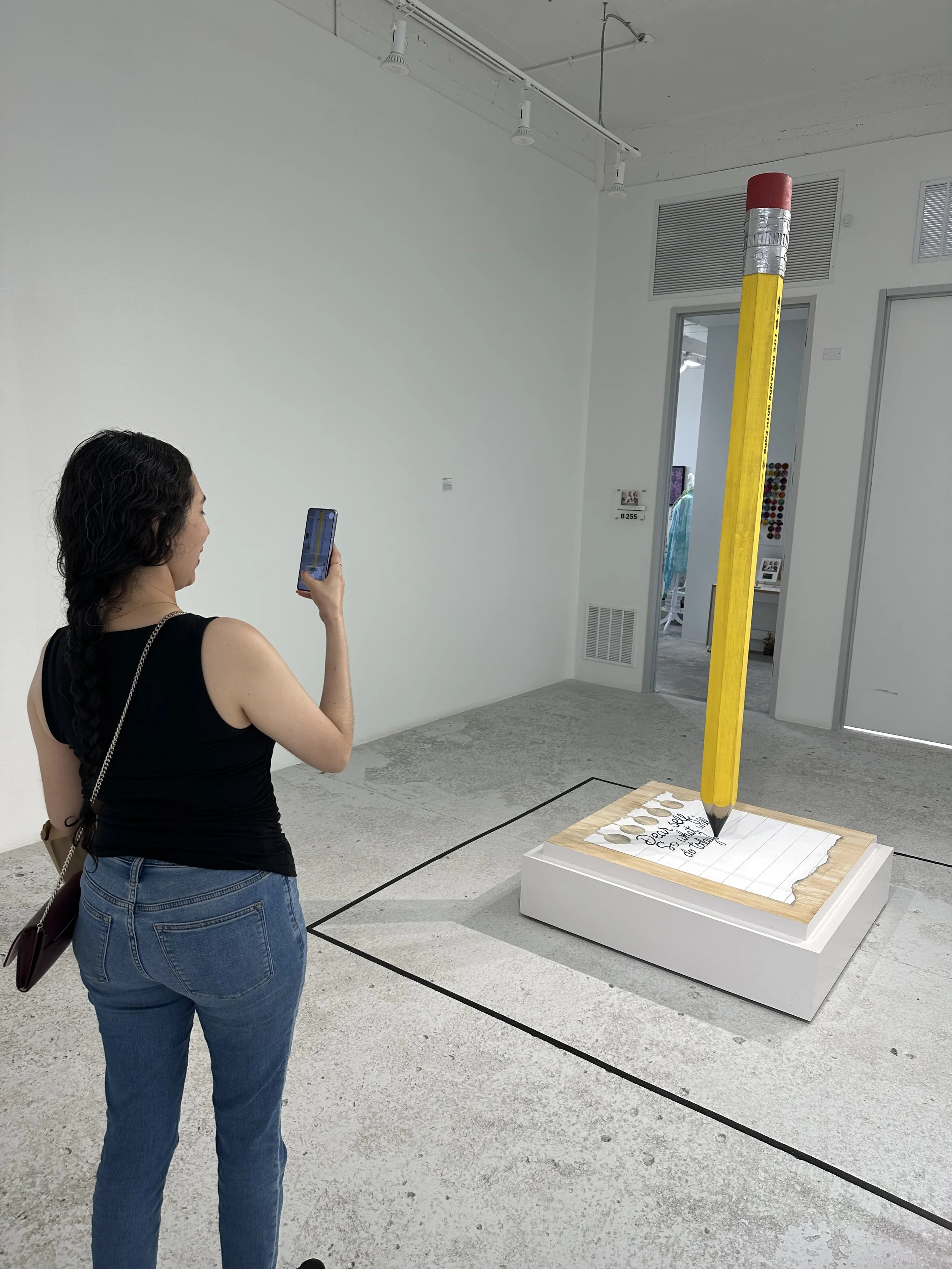 A woman taking a photo of a sculpture featuring a giant yellow pencil with a red eraser, standing upright on a platform with a piece of paper and coins. The sculpture is inside an art gallery with white walls and concrete floors.