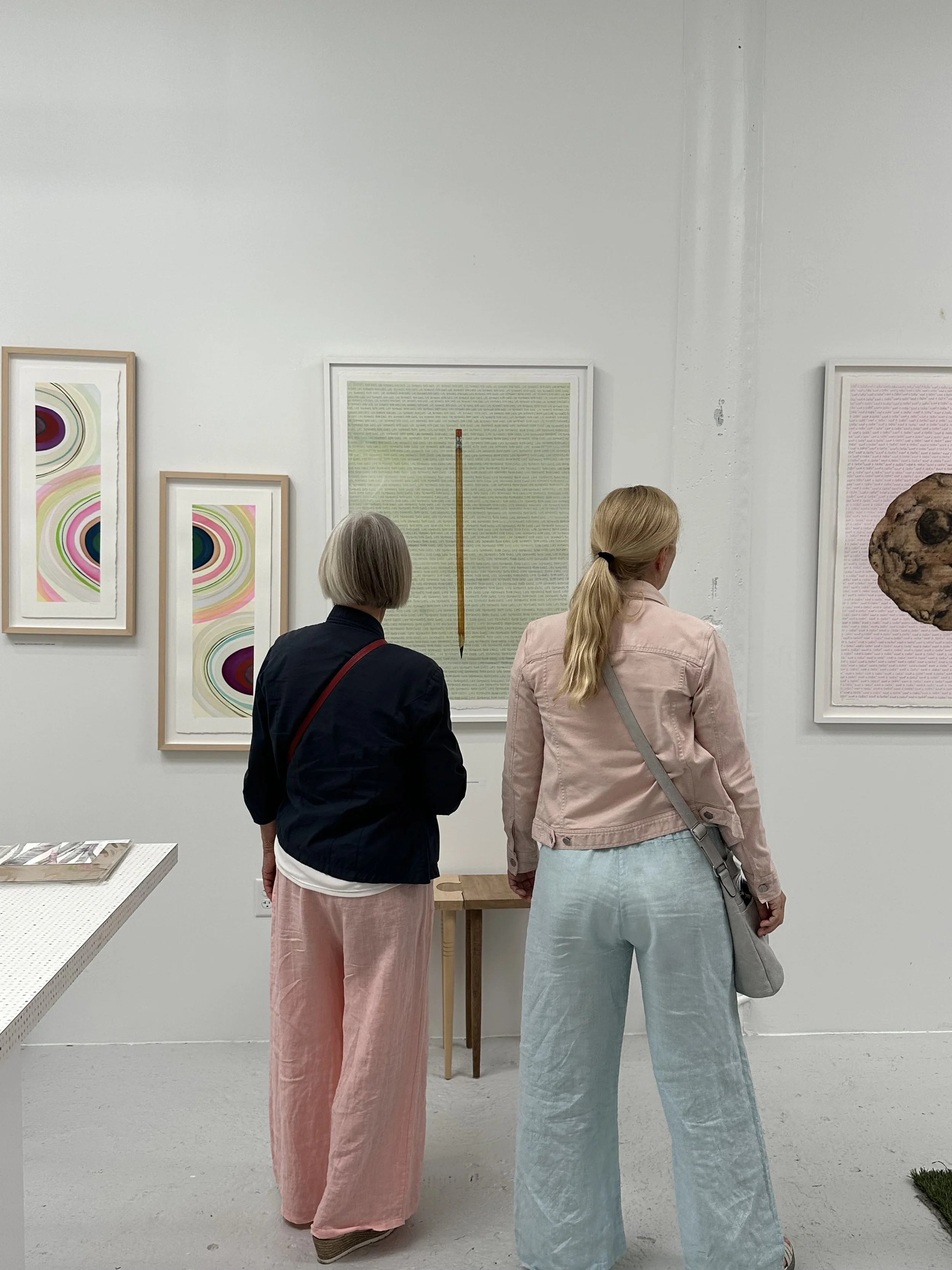 Two women viewing artwork in an art gallery, with framed abstract and text-based pieces on a white wall.