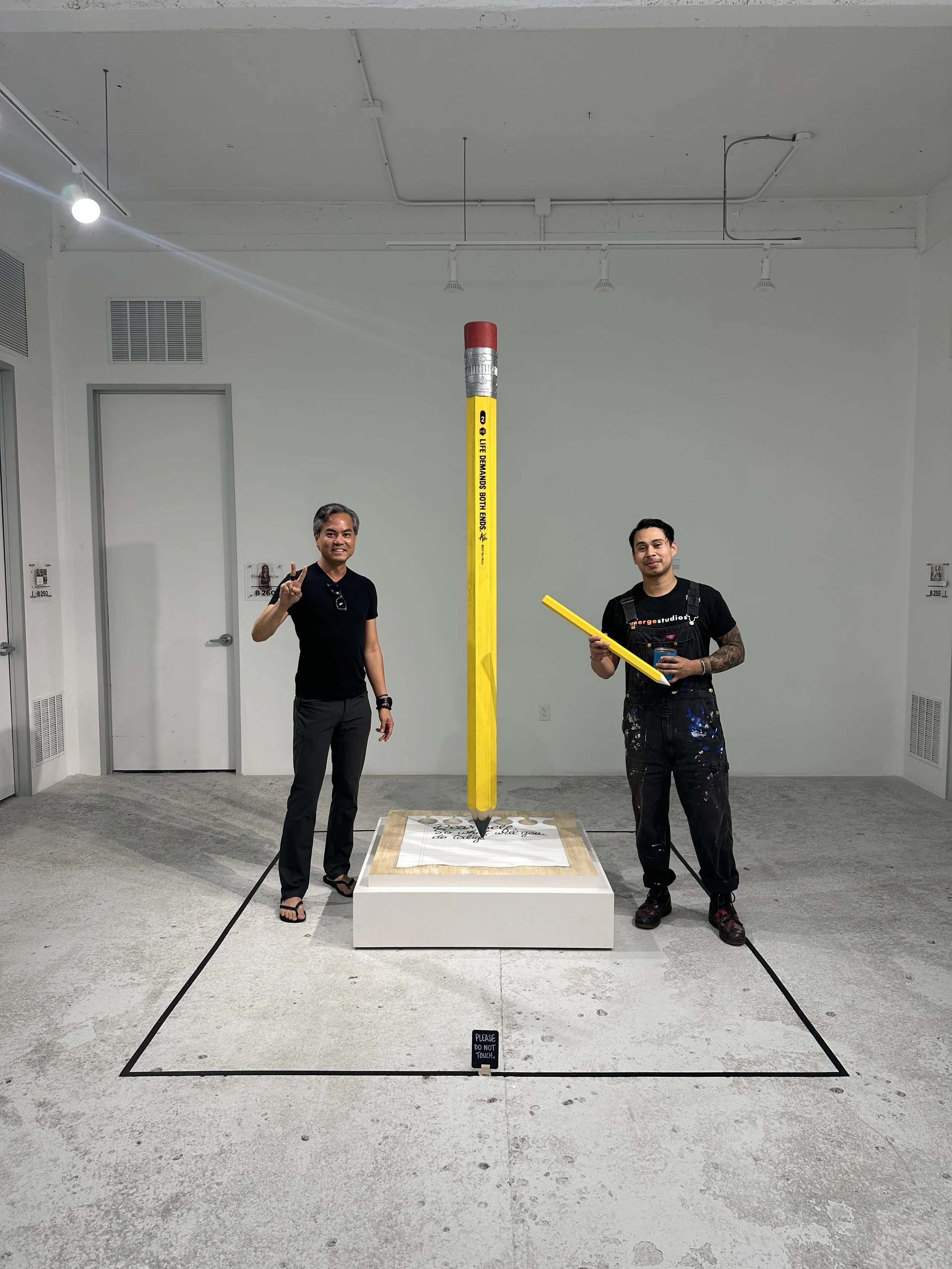 Two men standing next to an art installation of a large yellow pencil and a pencil sharpener inside a white gallery space. One man is making a peace sign, and the other is holding a smaller yellow pencil.
