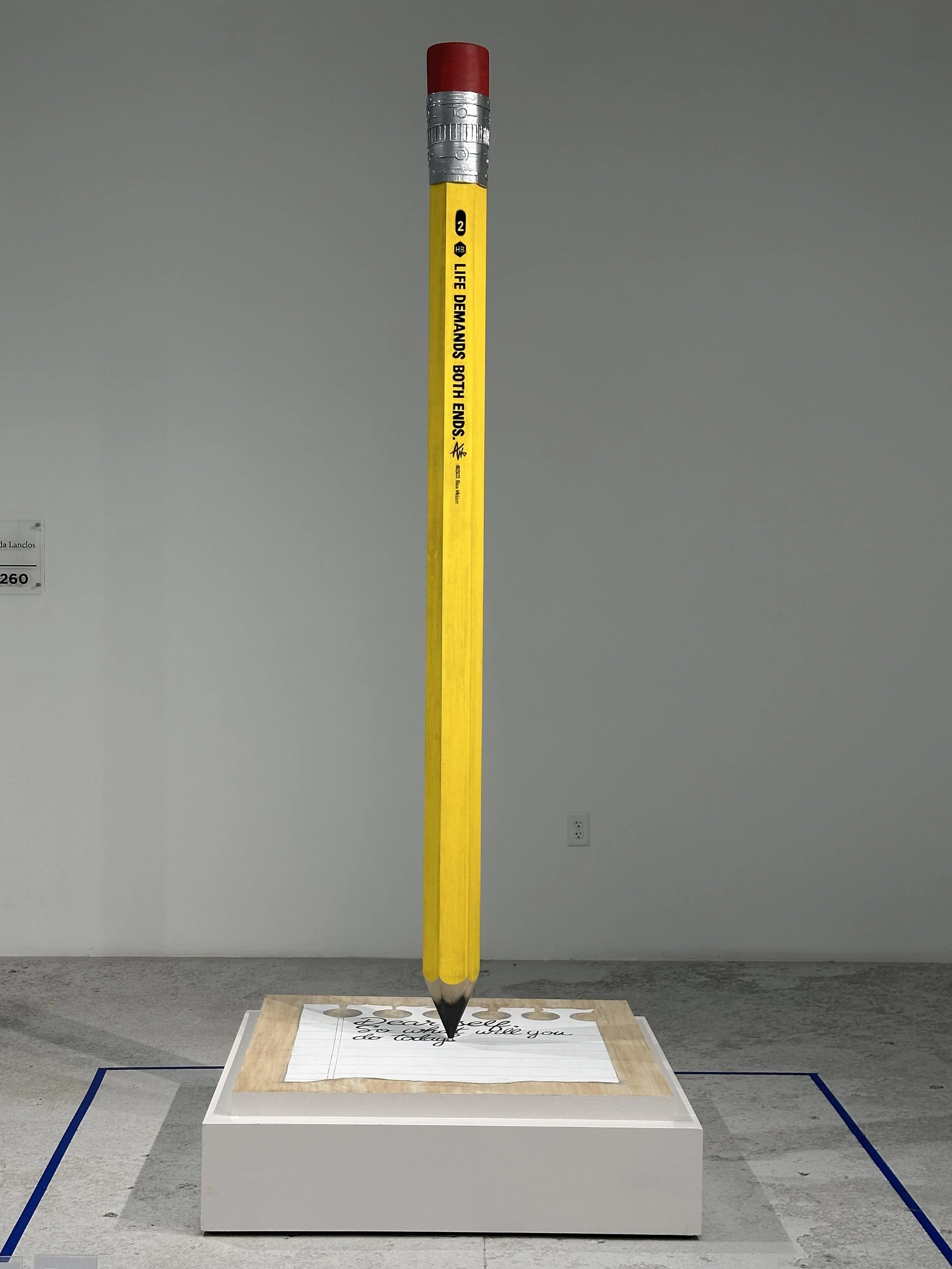 An art installation of a giant yellow pencil standing upright, with the pencil tip touching a framed piece of paper on a pedestal, inside a gallery space.