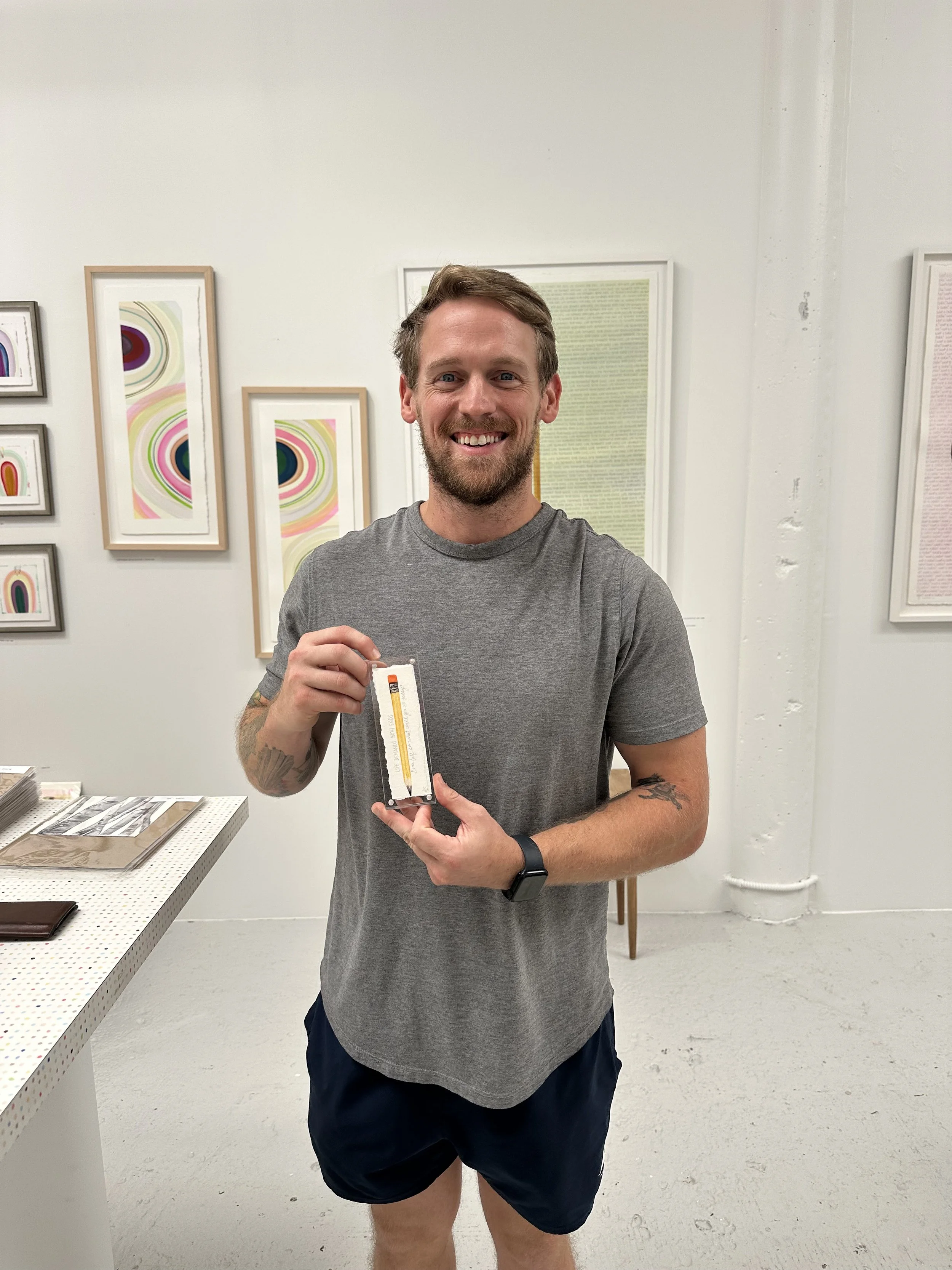 A smiling man with a beard and tattoos on his arms is holding a small object in an art gallery. He wears a gray T-shirt, black shorts, and a watch. The gallery walls are decorated with colorful abstract artwork.