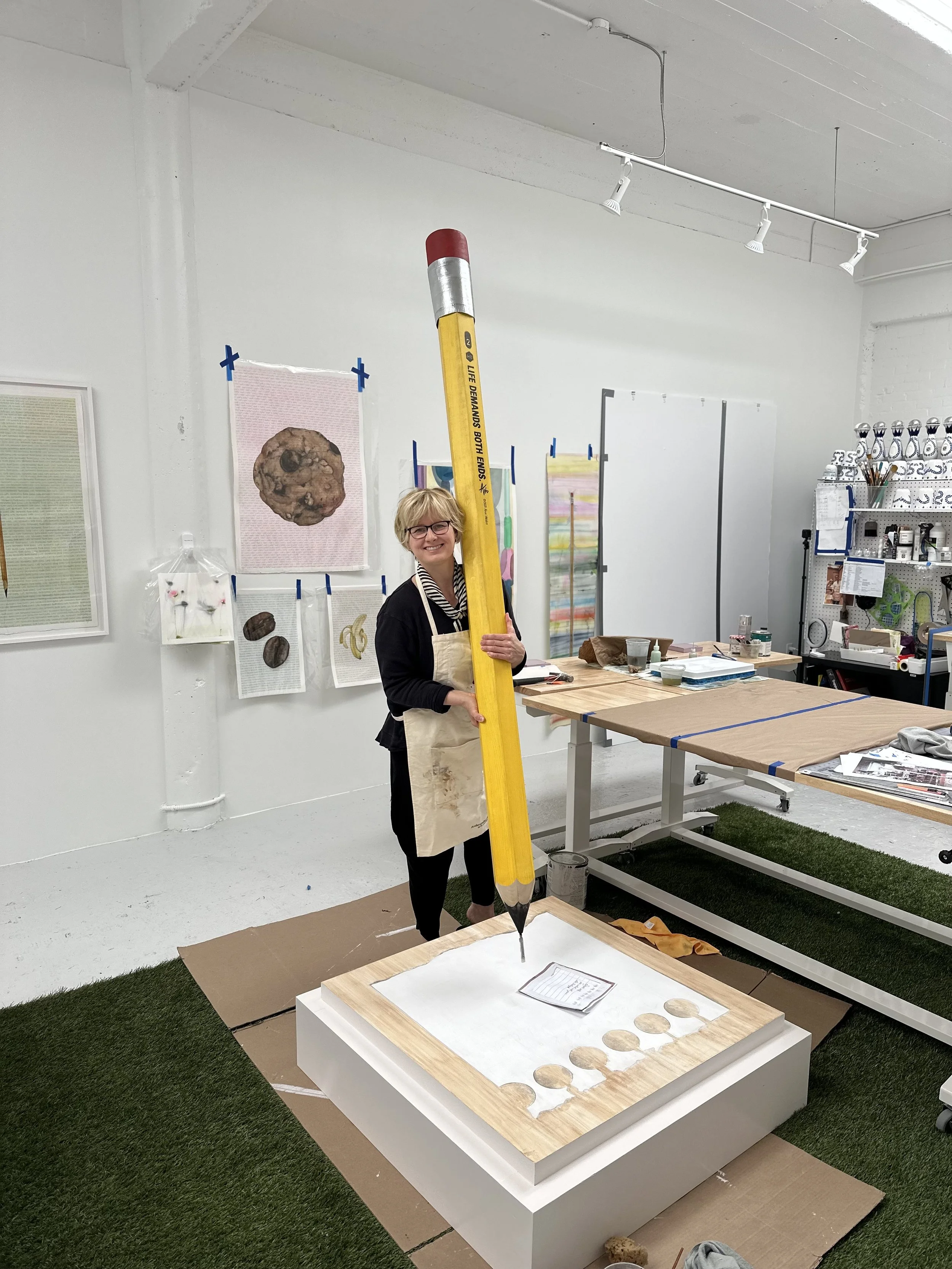 A woman wearing glasses and an apron is holding a large yellow pencil prop. She is standing in an art studio with paintings on the wall and art supplies around her.