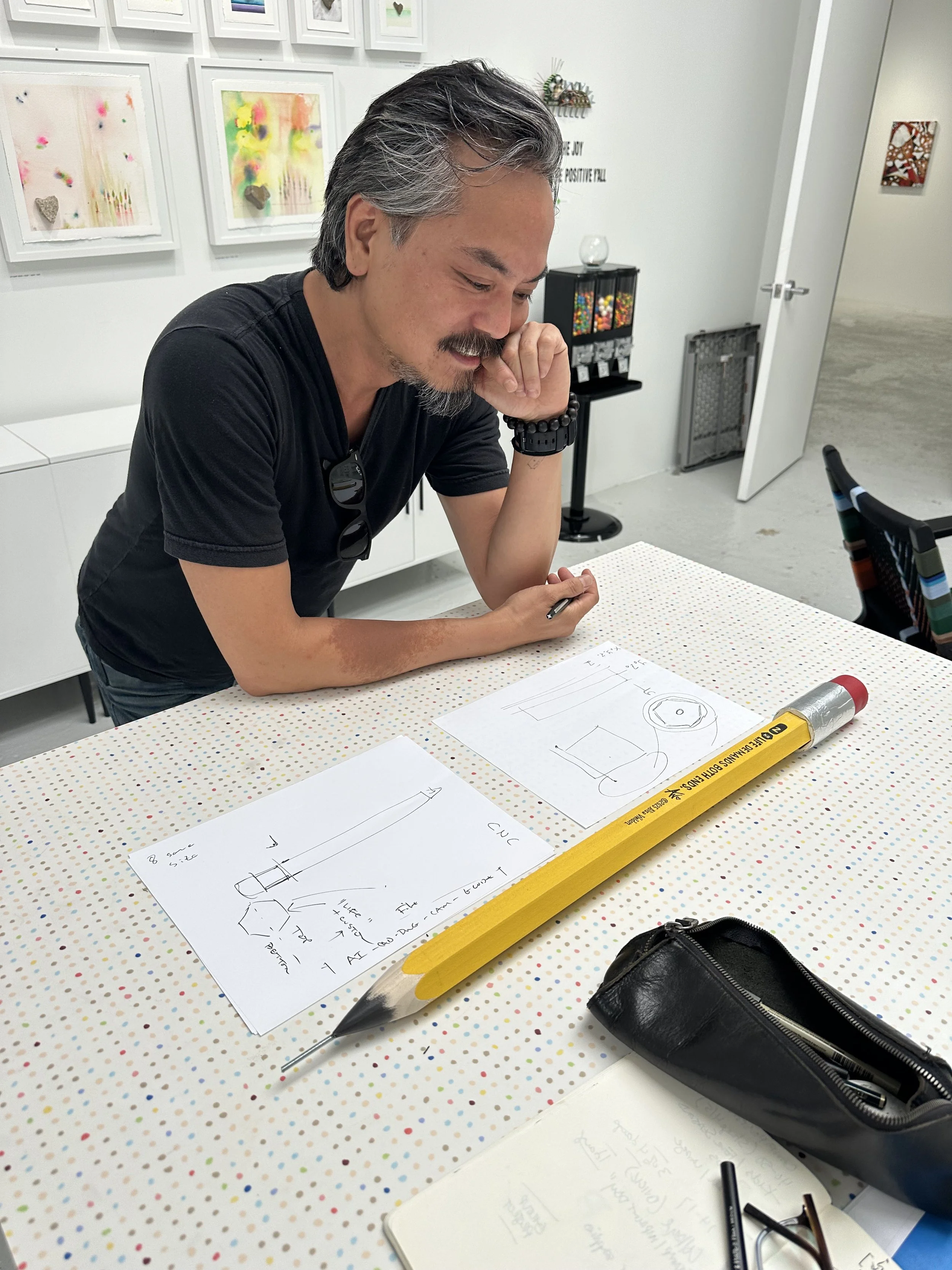A man with gray hair and a beard, wearing a black t-shirt, sitting at a table in an art gallery, looking at sketches and blueprints of a device, with a large yellow pencil and a black zippered bag on the table.